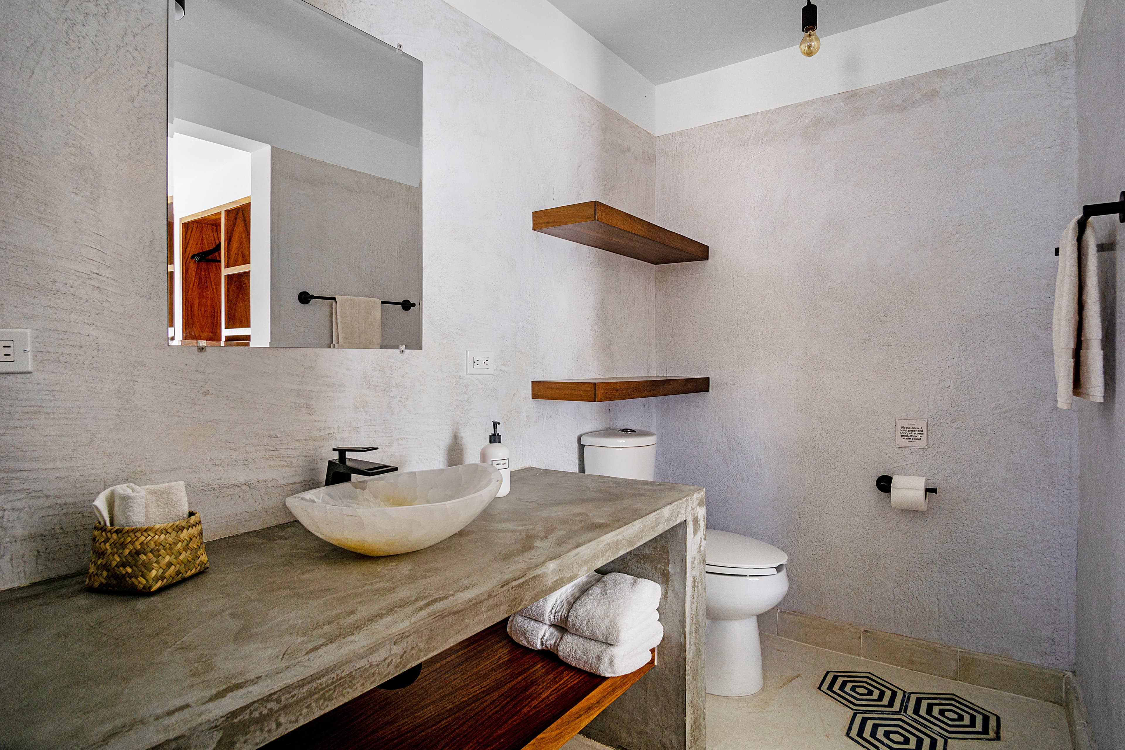 A modern bathroom featuring a concrete vanity, round sink, wooden shelves, and minimalist decor.