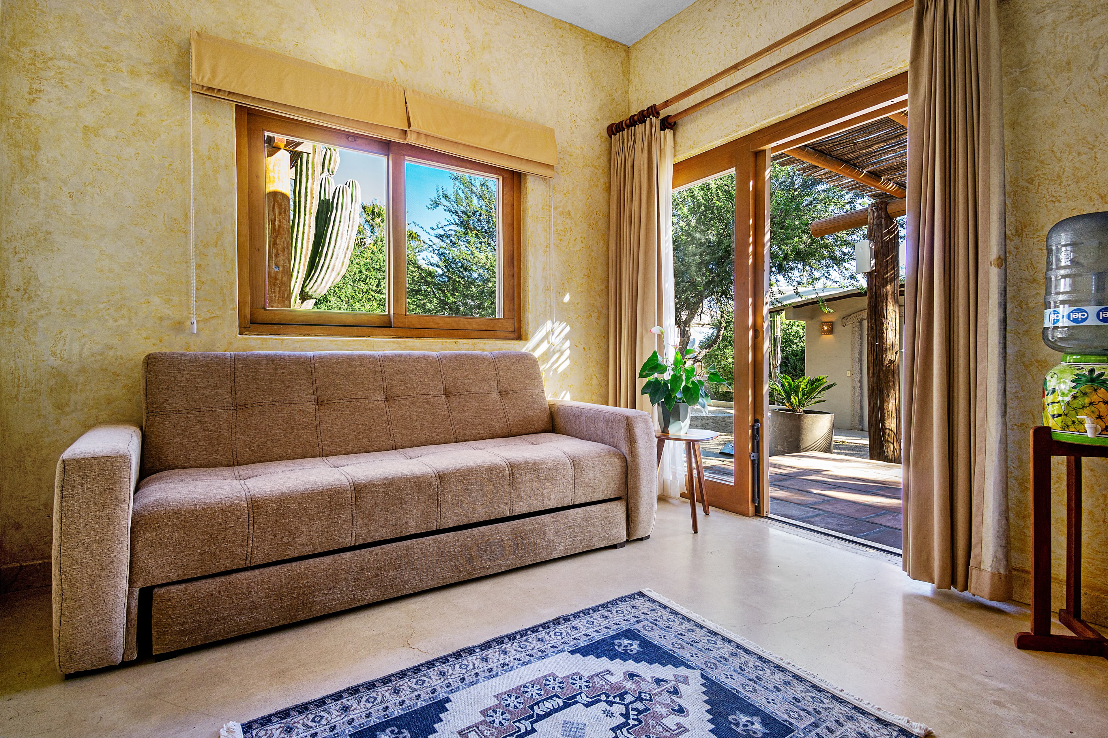 Cozy living room featuring a beige sofa, a patterned rug, a large window with a cactus view, and a patio door.