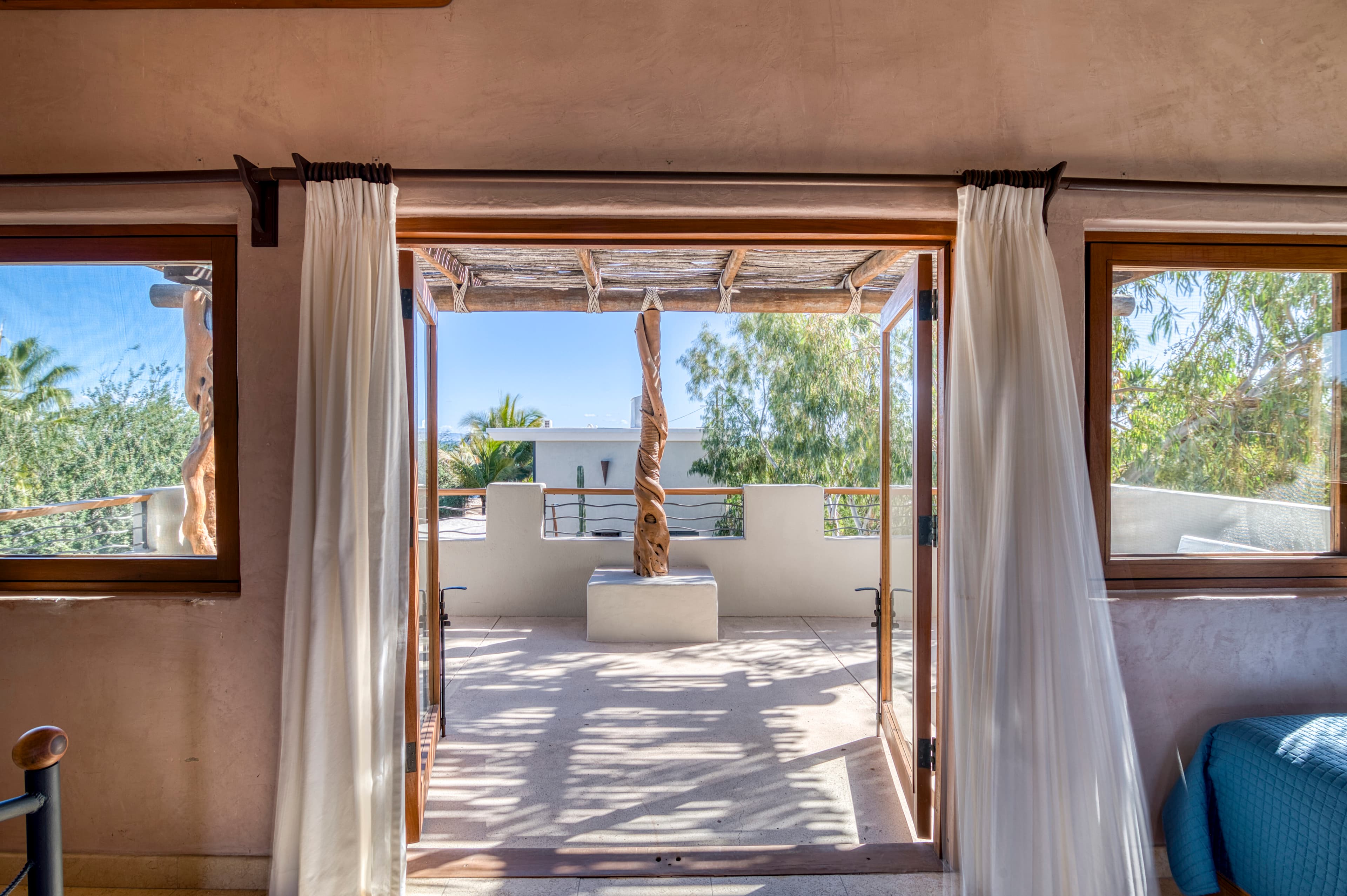 A view from an open doorway leading to a sunlit terrace with a decorative pillar and lush greenery beyond.