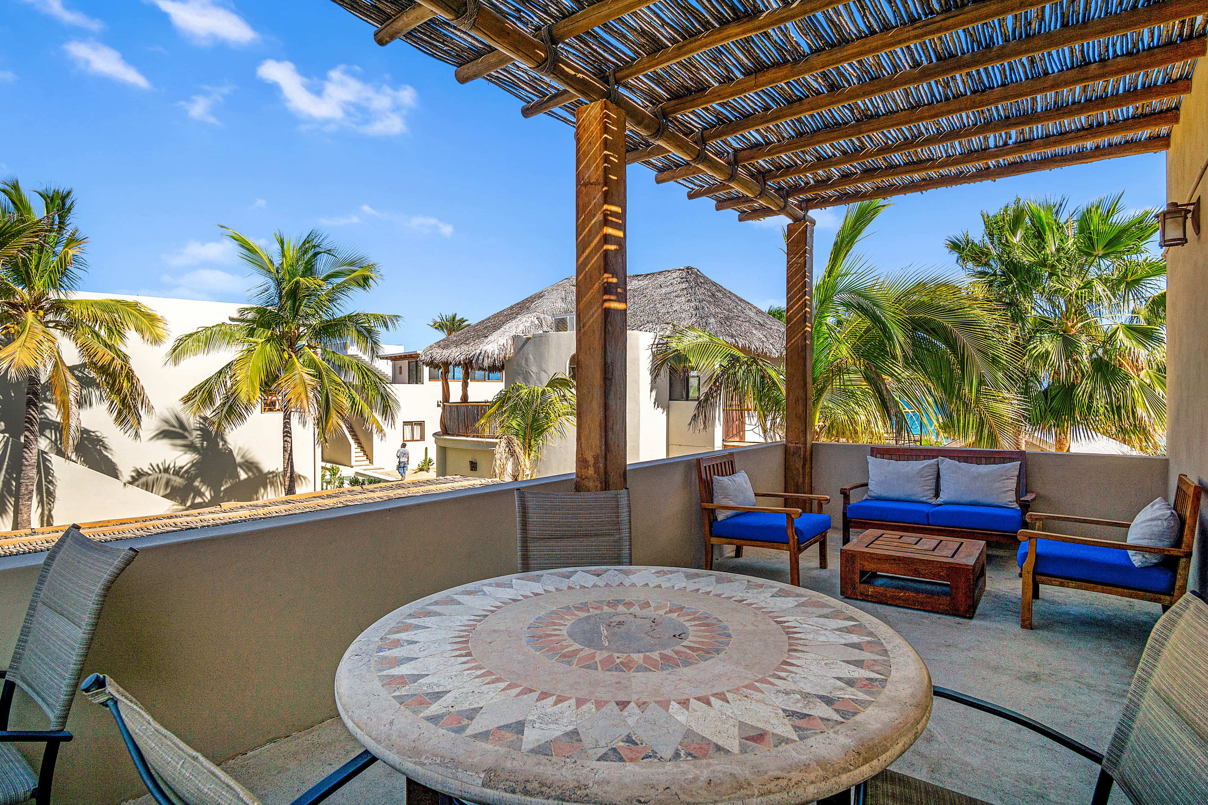 A sunlit balcony with a round mosaic table and comfortable seating, surrounded by palm trees and tropical scenery.
