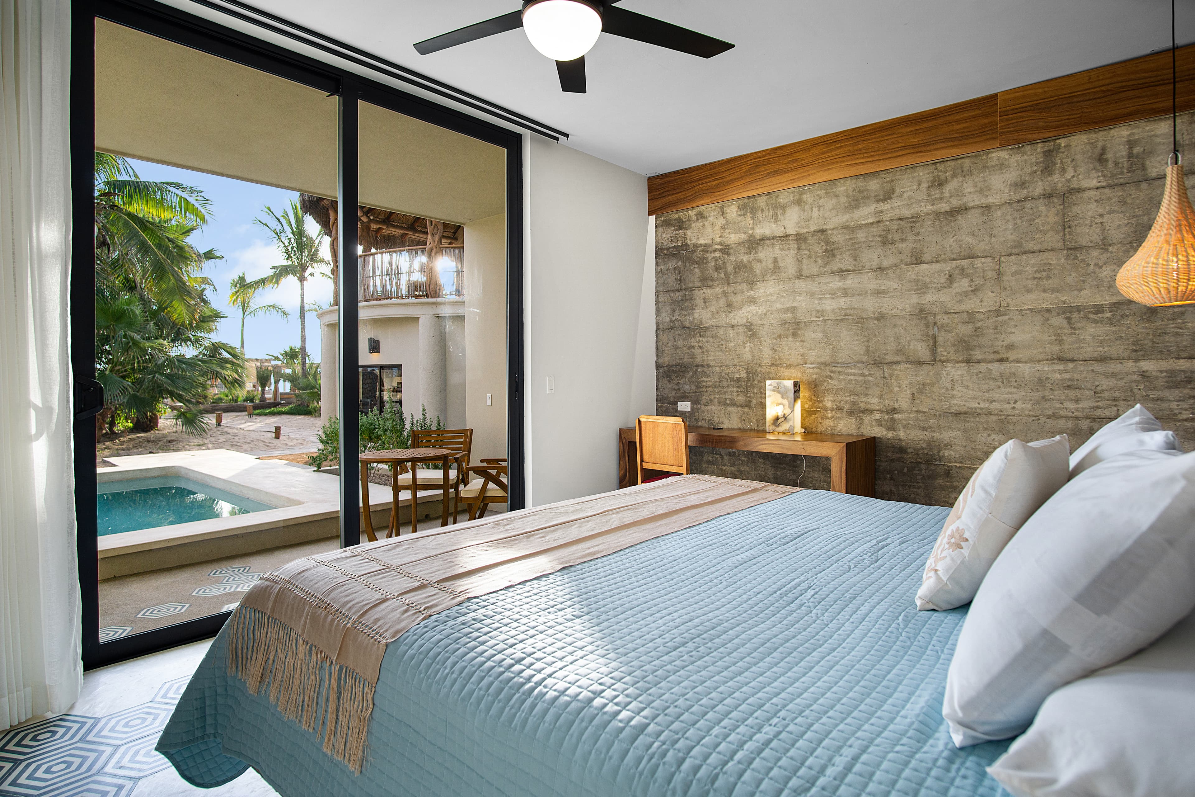 A stylish bedroom with a large window overlooking a tropical pool and patio area.
