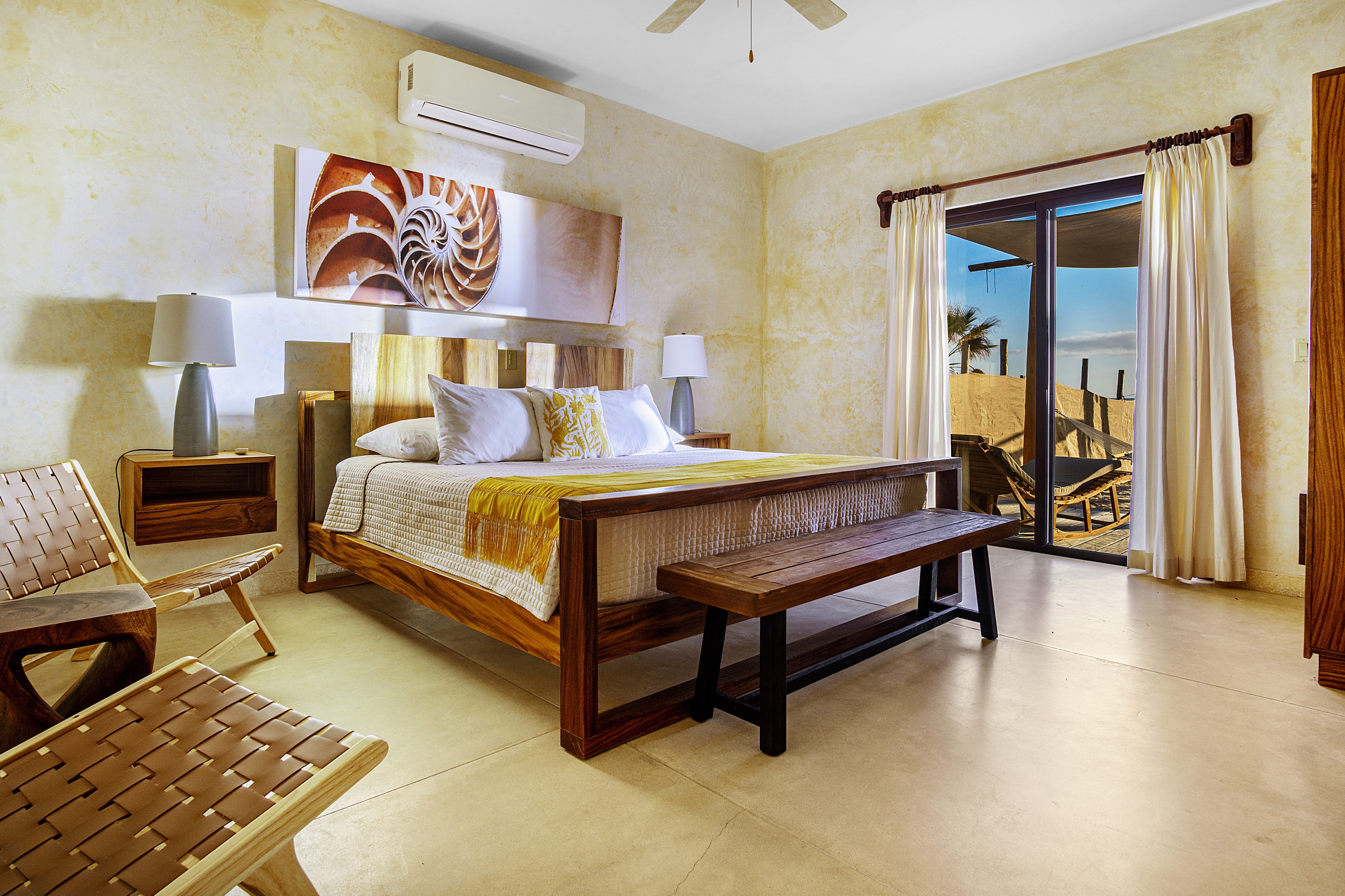 A cozy bedroom with a wooden bed, two lamps, a bench, and a view of a patio outside through sliding glass doors.