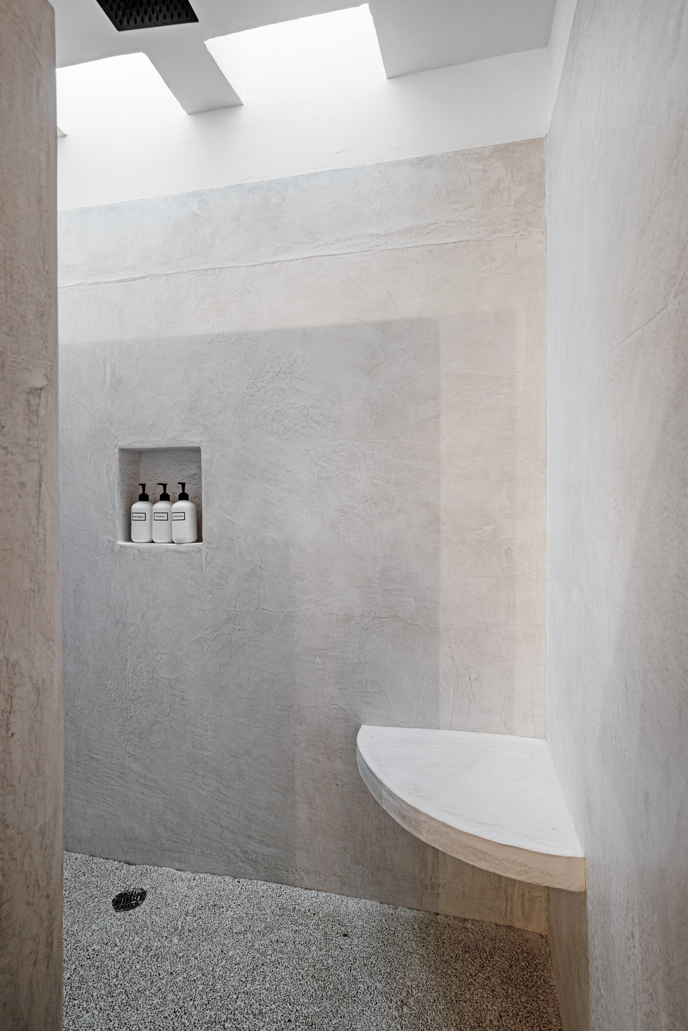 A minimalist shower with smooth concrete walls, a bench, and a shelf holding three bottles.