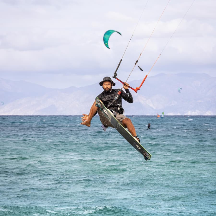 A kitesurfer performs a jump over the ocean with colorful kites in the background.