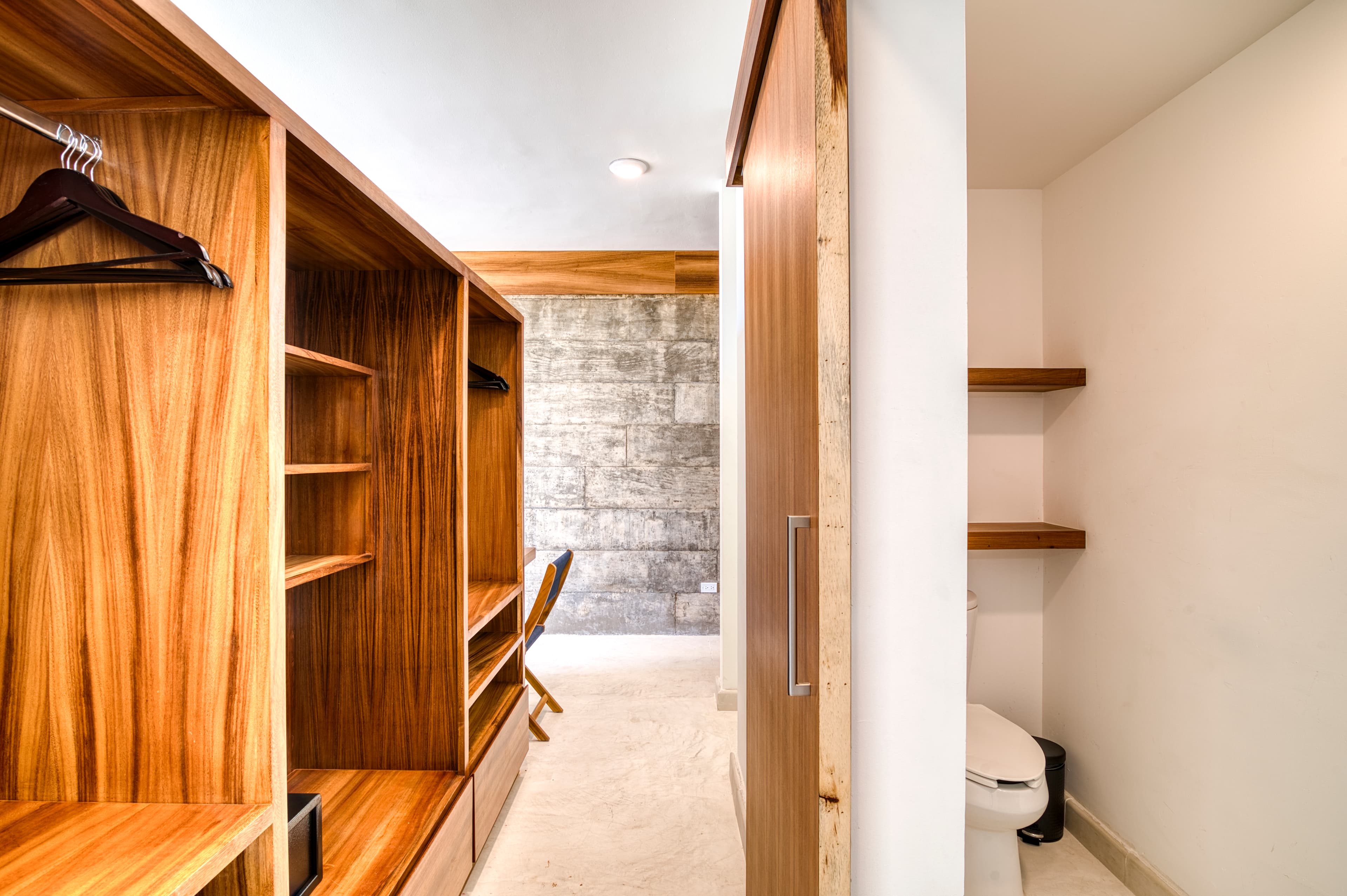 A modern, minimalistic closet space featuring wooden shelves, a chair, and an adjacent bathroom area.