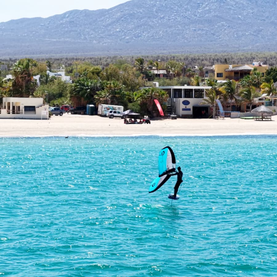 A person on a hydrofoil board glides over turquoise waters near a sandy beach and mountains.