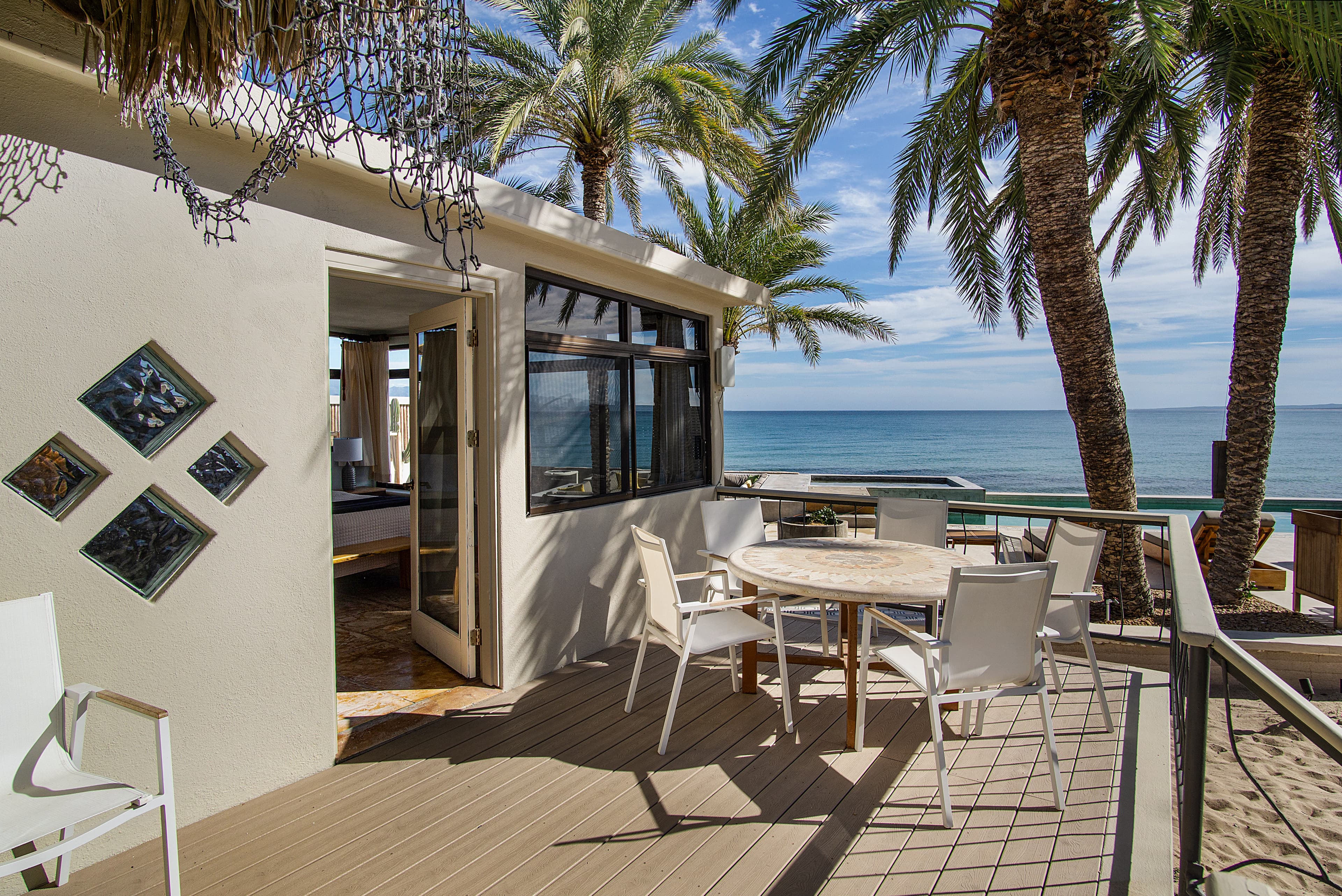 A cozy outdoor patio with a table and chairs overlooks the ocean, surrounded by palm trees and a beachside home.