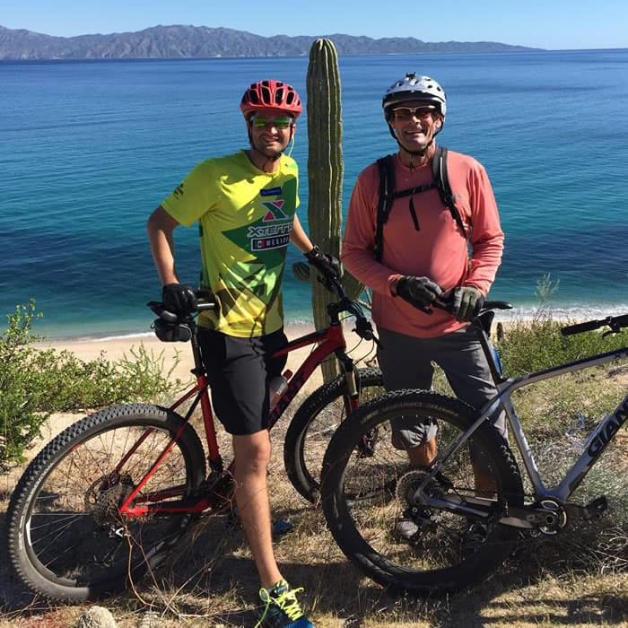 Two men with mountain bikes posing by a cactus by the shore, with a blue ocean and mountains in the background.