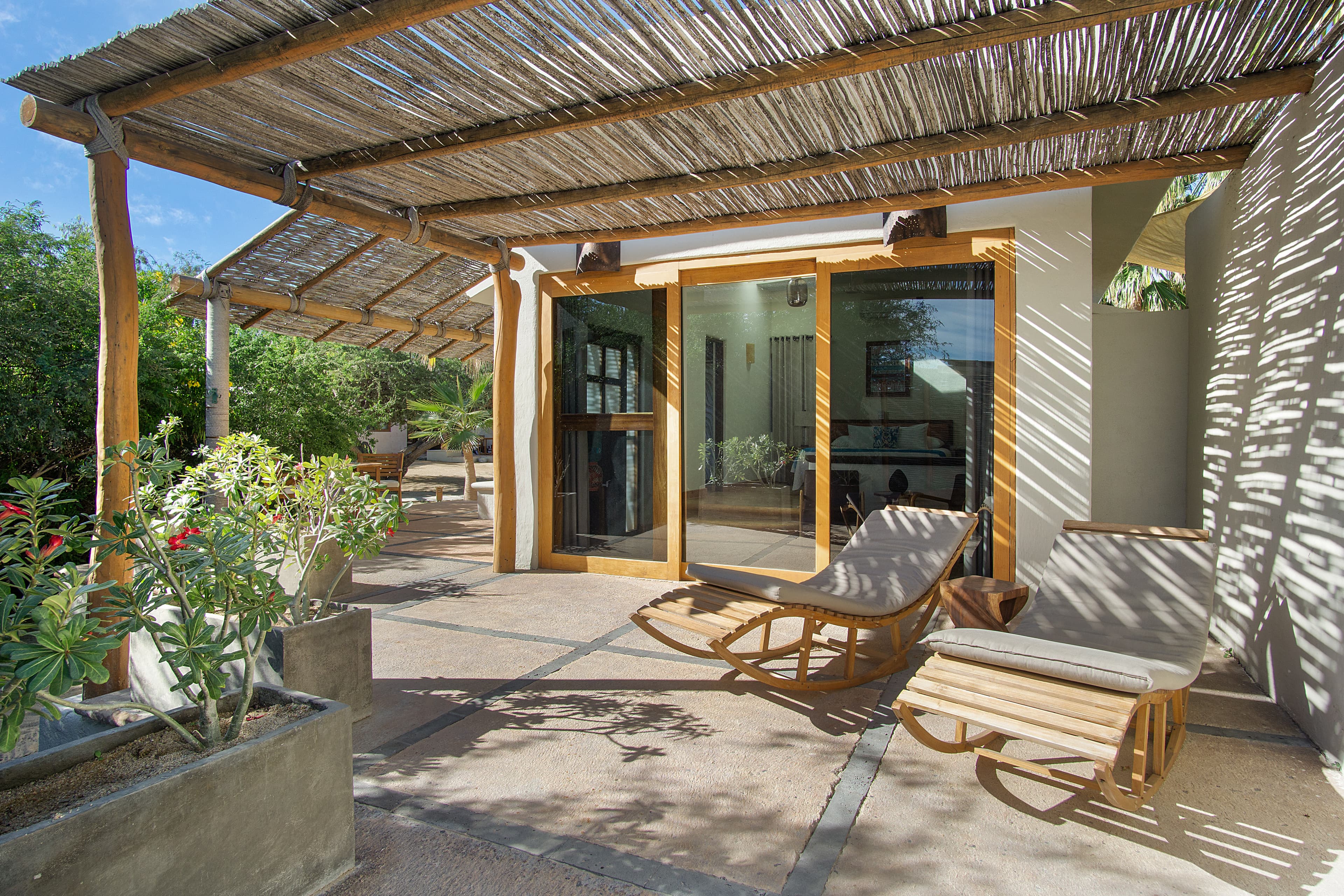 A cozy outdoor seating area with two lounge chairs beneath a thatched roof and surrounded by plants.