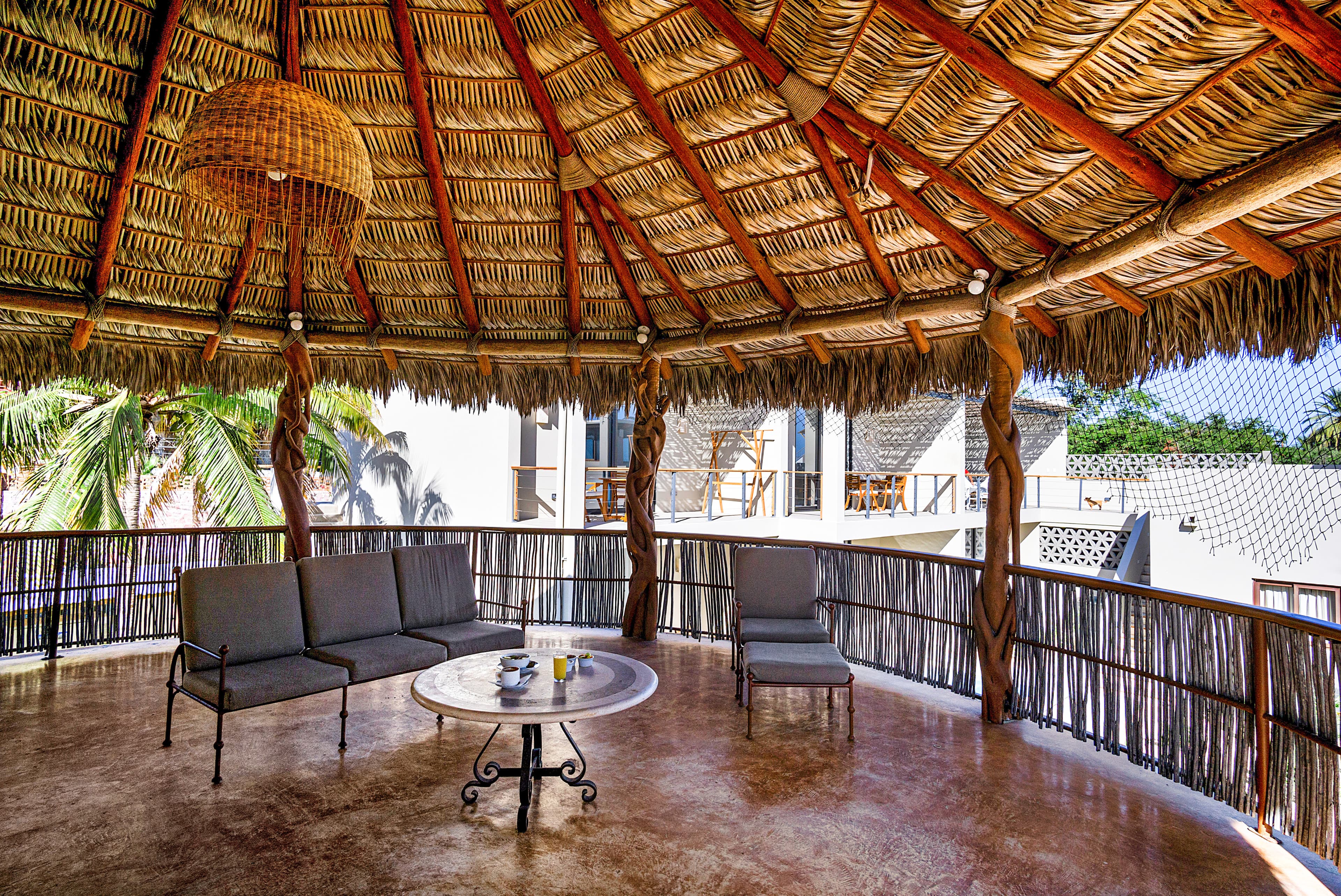 A rustic outdoor seating area with a thatched roof, featuring a round table and two chairs surrounded by tropical plants.
