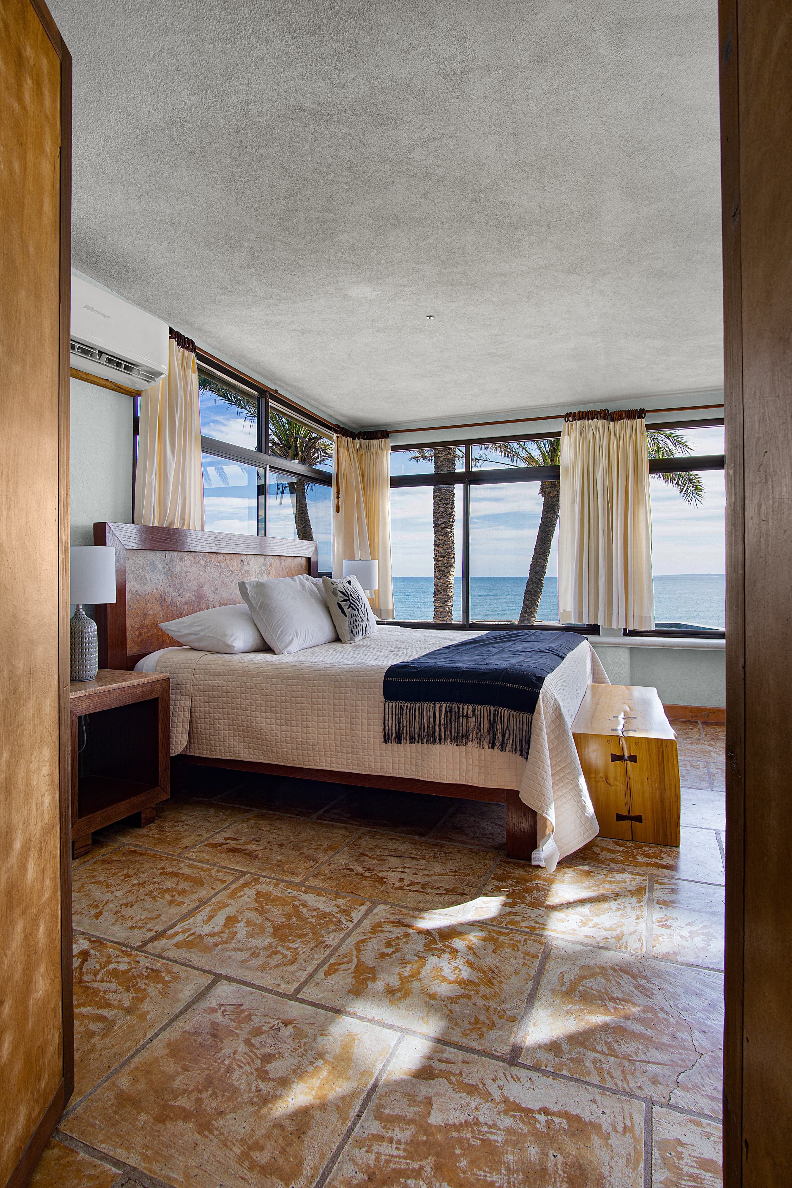 A serene bedroom with a large window view of the ocean and palm trees.