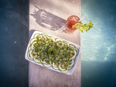A platter of cucumber slices garnished with herbs beside a cocktail in a glass.