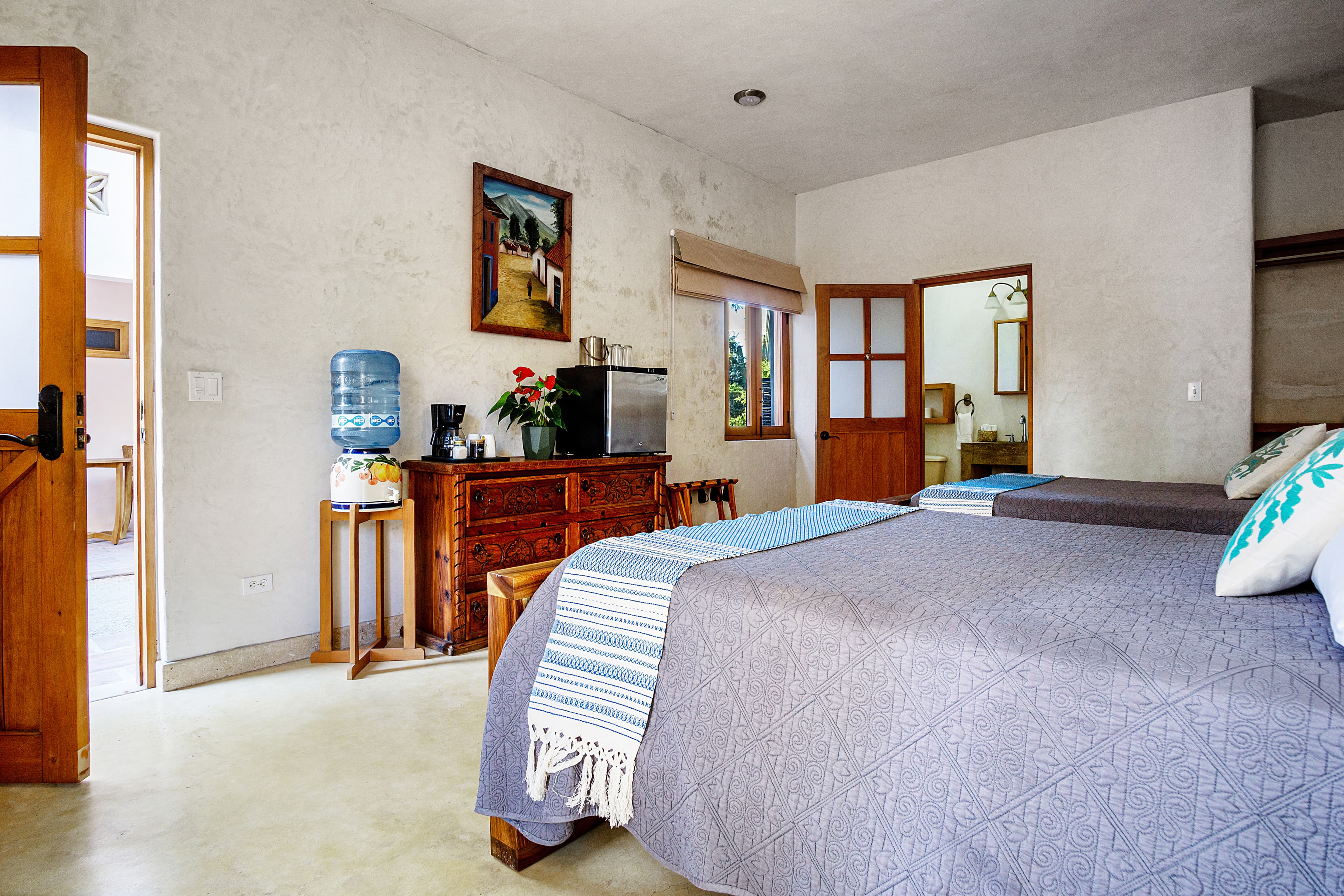 A cozy, minimalistic bedroom featuring two beds, a wooden dresser, and an open door leading to an outdoor space.