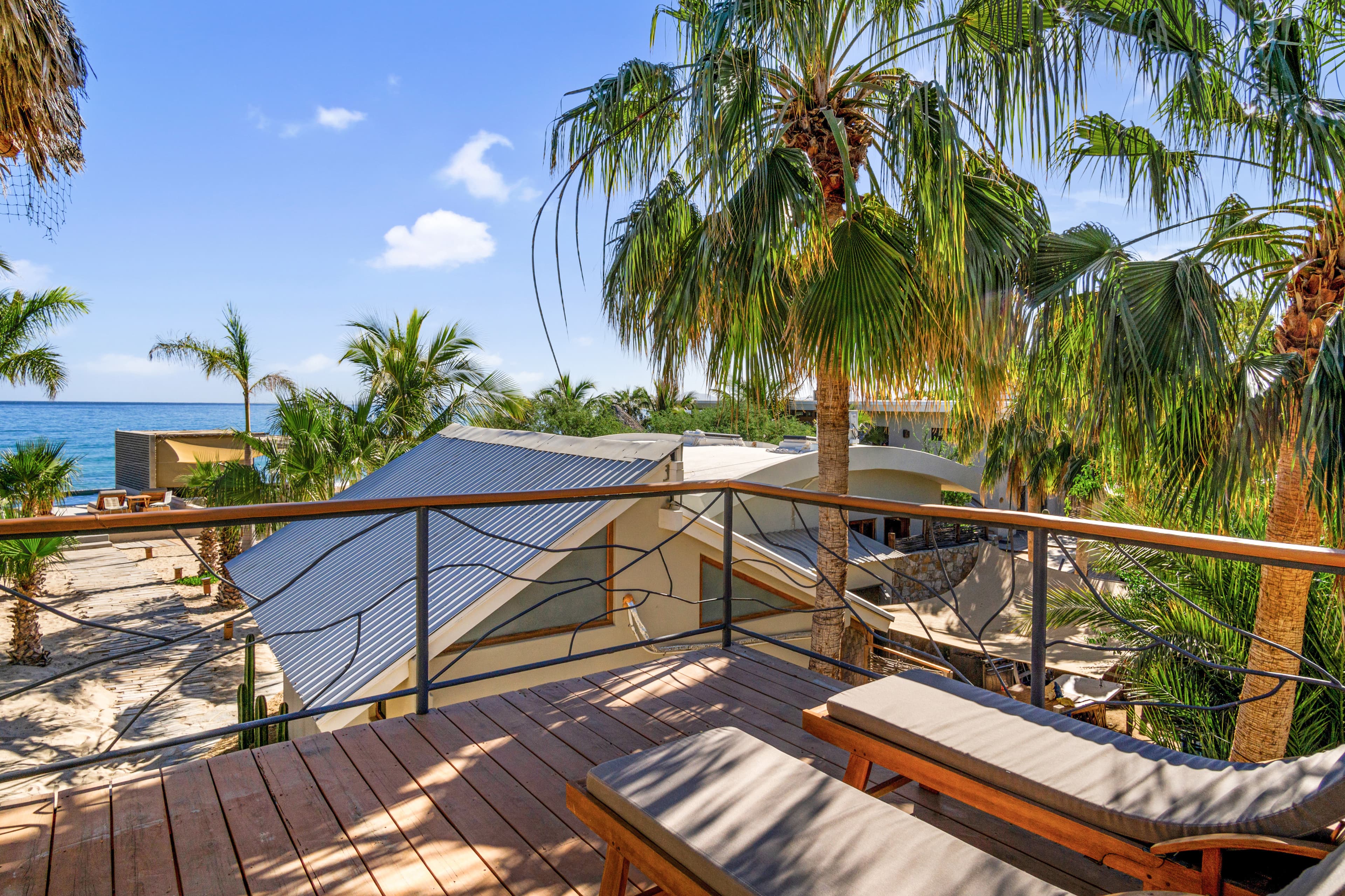 View from a wooden deck overlooking a beach with palm trees and the ocean in the background.