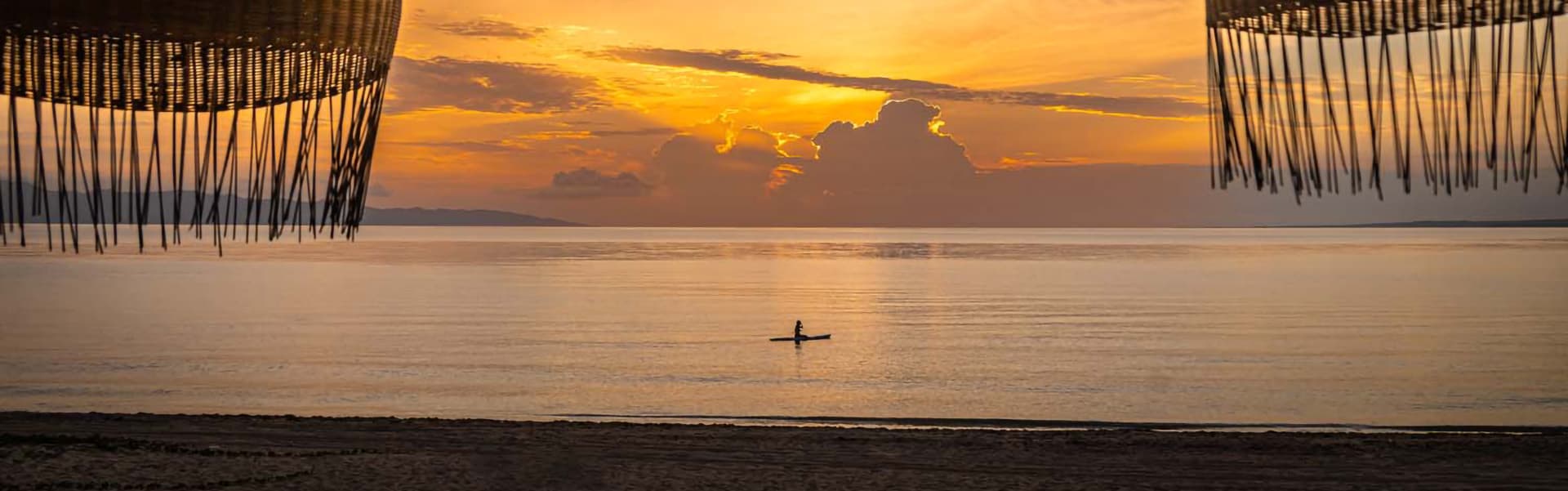A serene sunset over calm waters, framed by woven beach decor, with a lone kayaker in the distance.