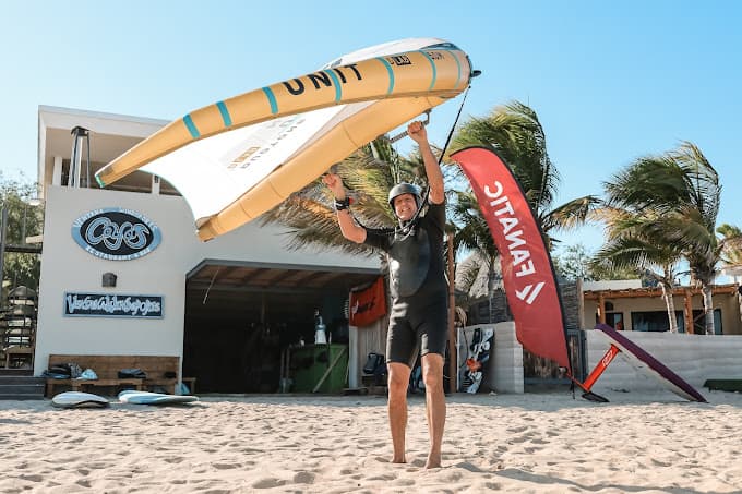 A person in a wetsuit holds up a wing on a beach with surfboards and palm trees in the background.