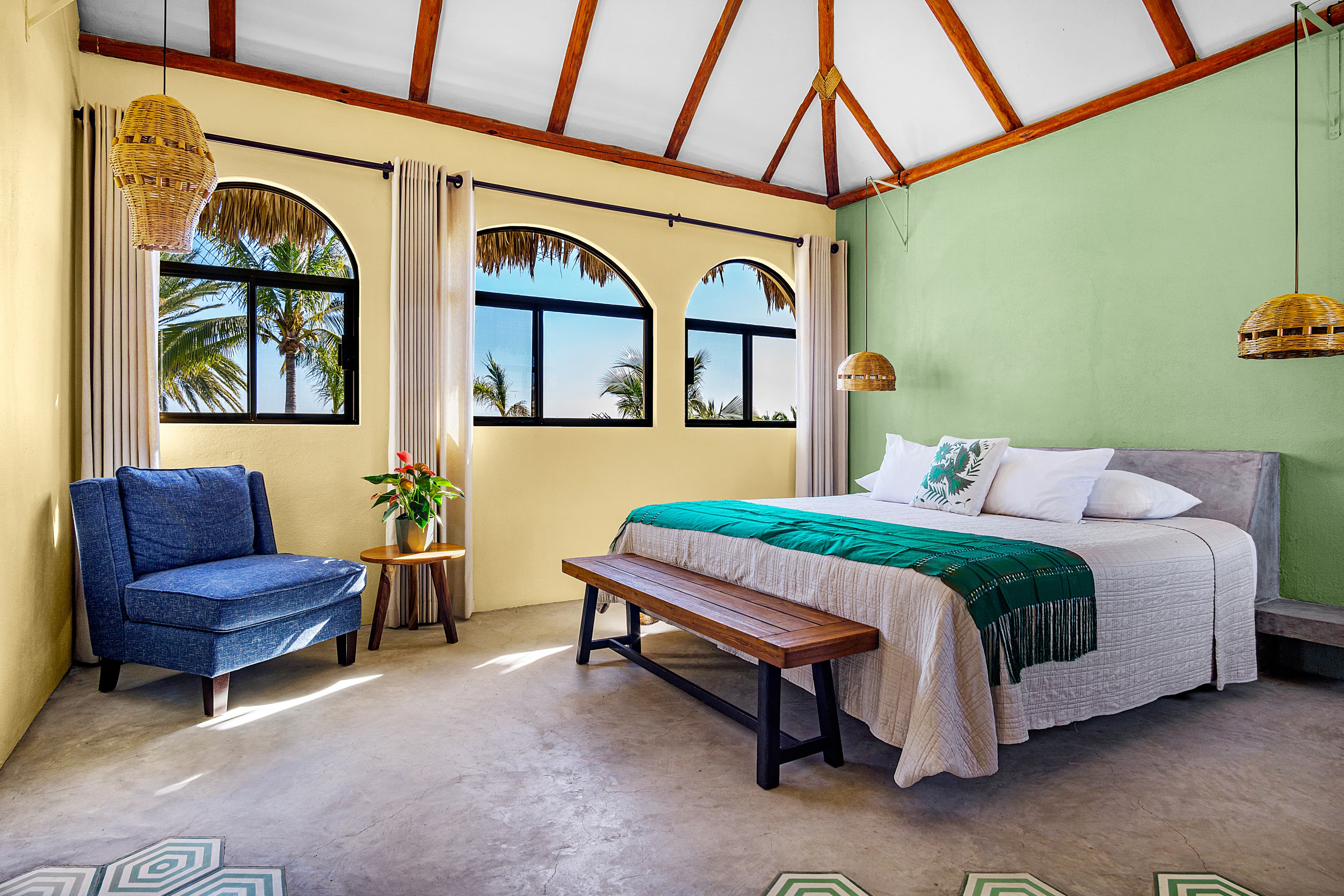 Bright, airy bedroom with green walls, a cozy bed, a lounge chair, and large windows showcasing palm trees outside.