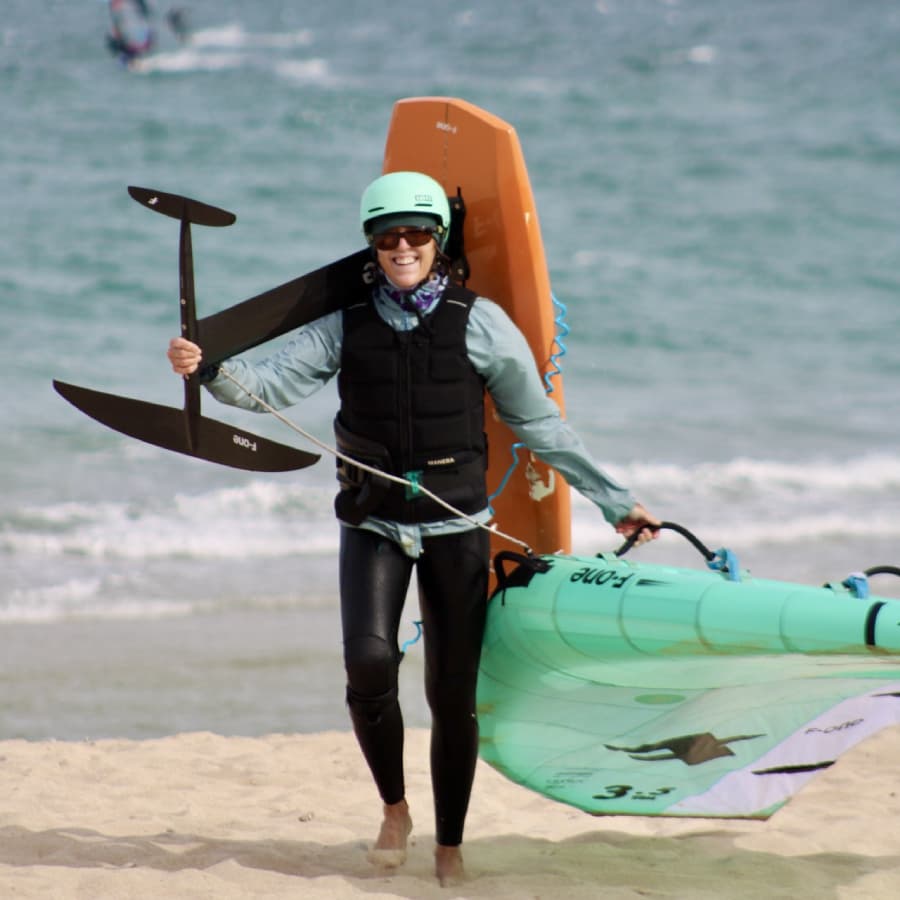 A smiling person in a helmet carries a foil board and wing on a beach.