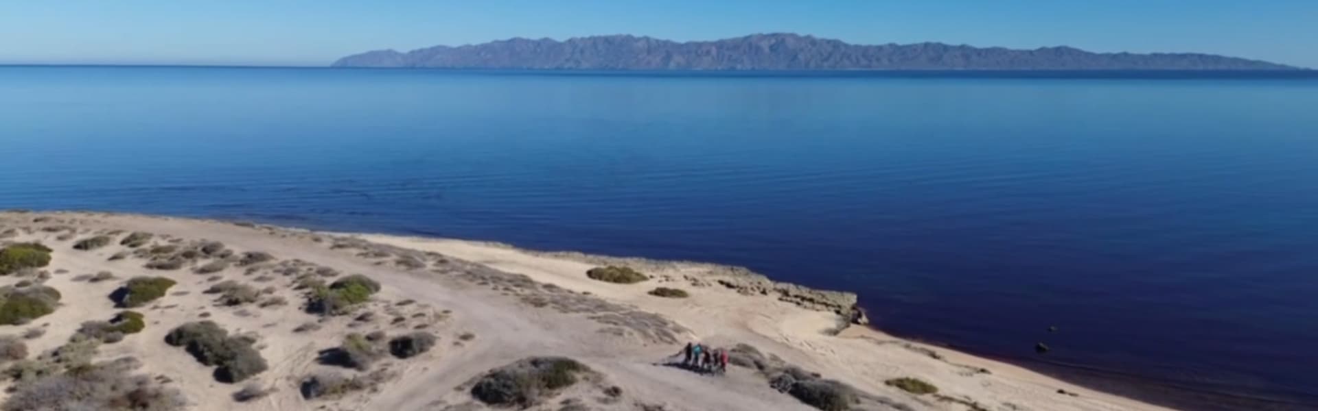 Aerial view of a sandy beach with a group of people near the water, and distant mountains in the background.
