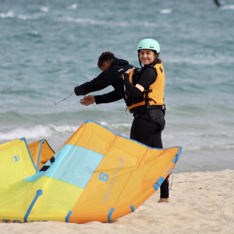 A woman in a wetsuit and helmet smiles while preparing a colorful kite on the beach.