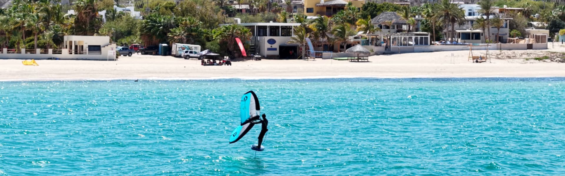 A kite surfer glides over turquoise ocean waters near a sandy beach and palm trees.