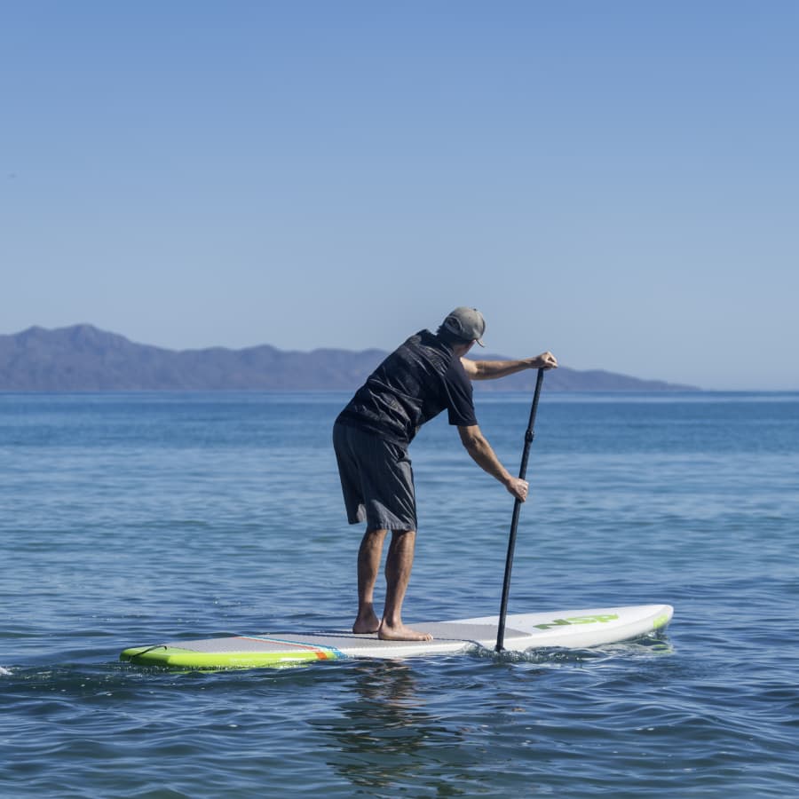 A person paddleboards on calm water with mountains in the background.