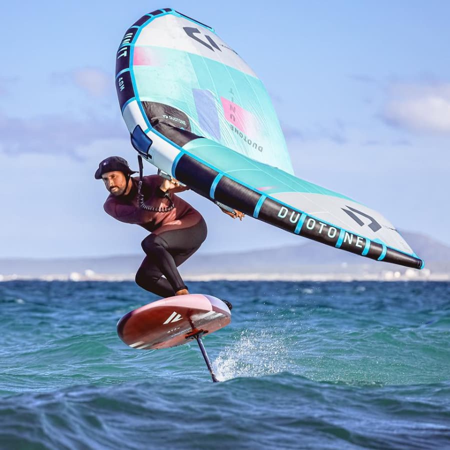 A person performs a foiling maneuver on a surfboard while holding a colorful wing in the ocean.