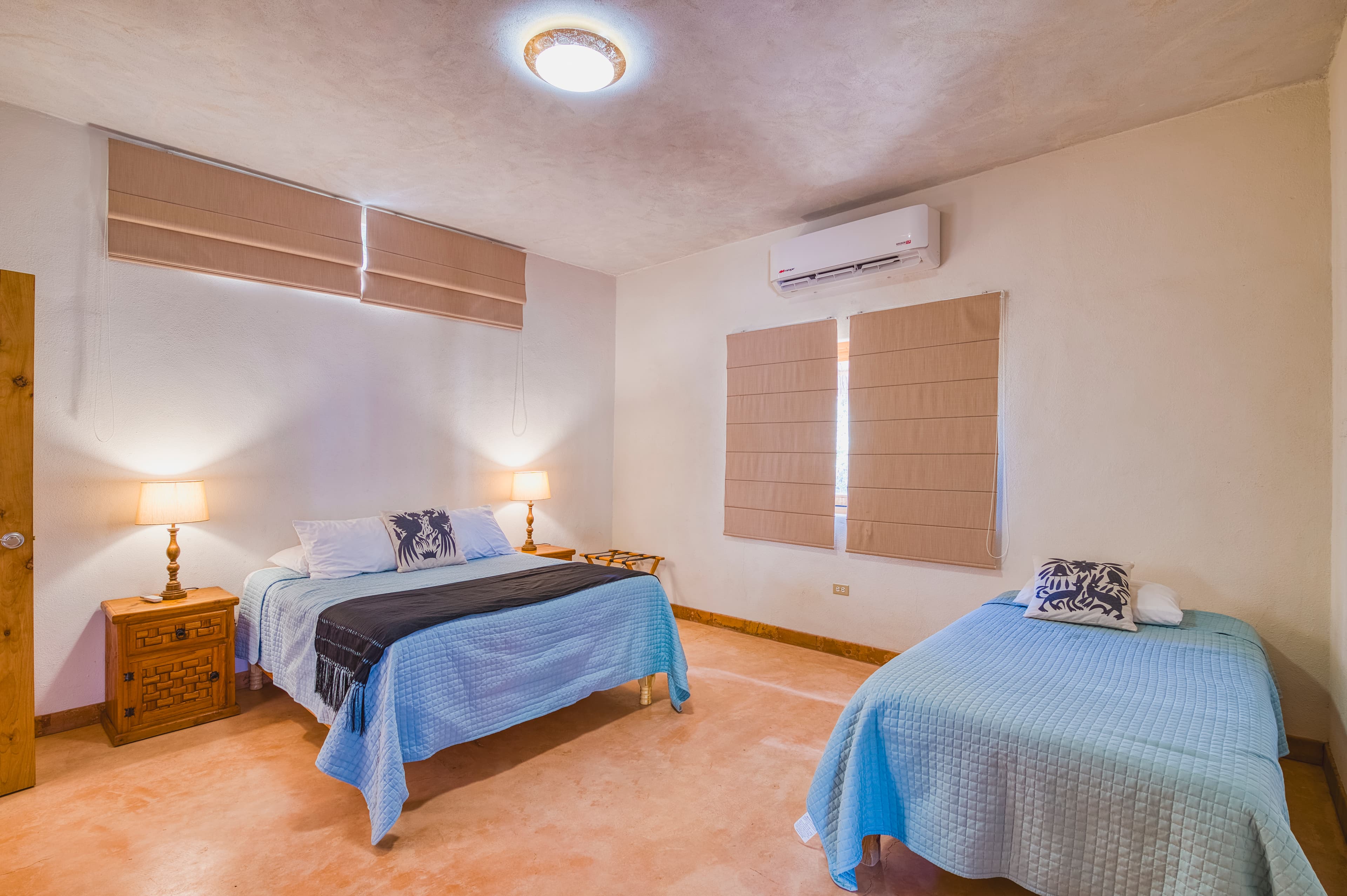 A bright bedroom featuring two beds with blue blankets, lamps on bedside tables, and horizontal blinds on the windows.