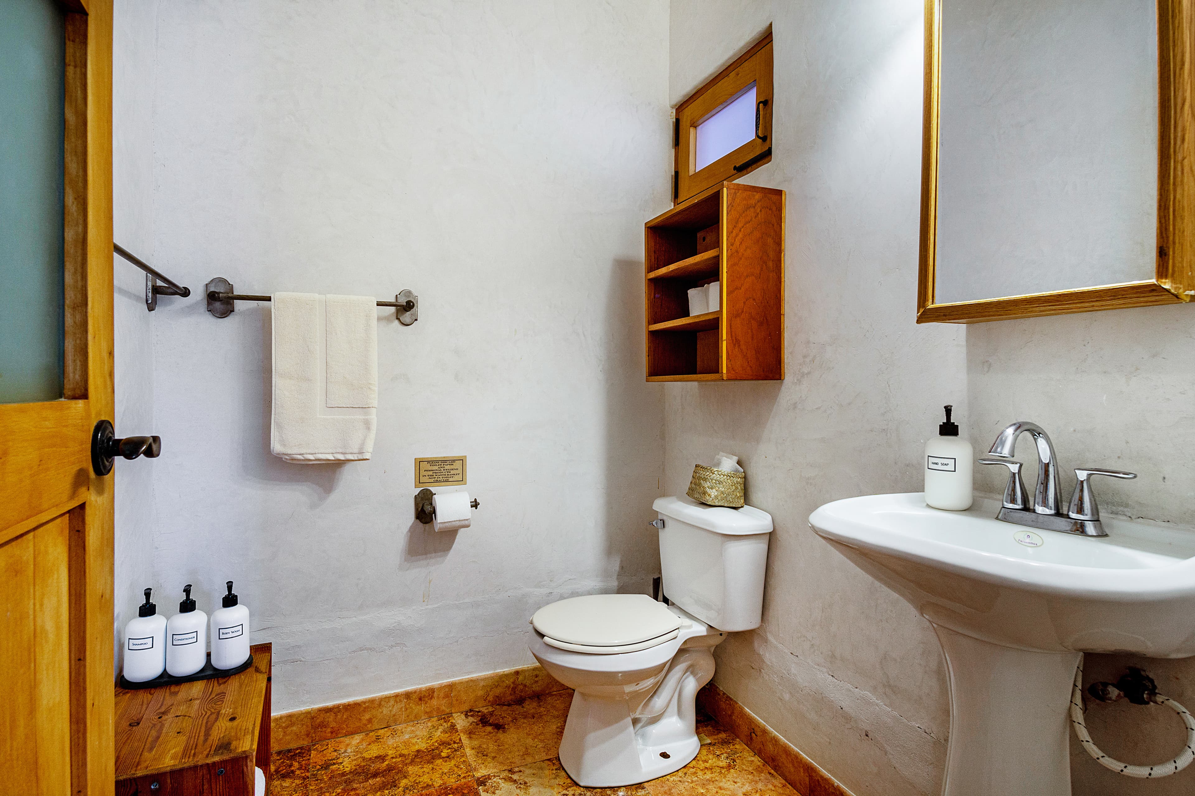 A modern bathroom featuring a toilet, sink, mirror, and wooden shelving with toiletries.