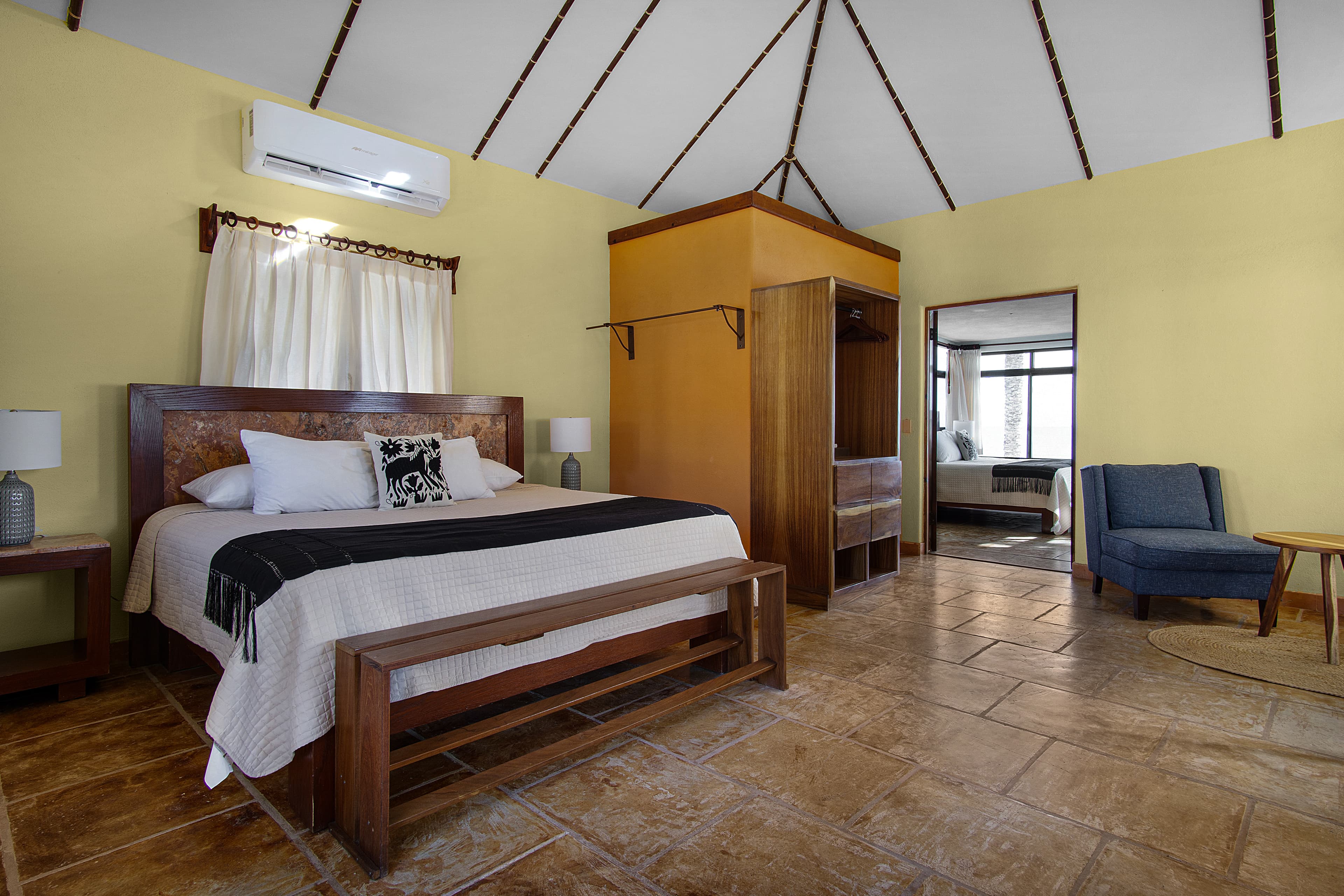 A cozy bedroom with a wooden bed, two lamps, a wardrobe, and a seating area, featuring warm yellow walls and a high ceiling.