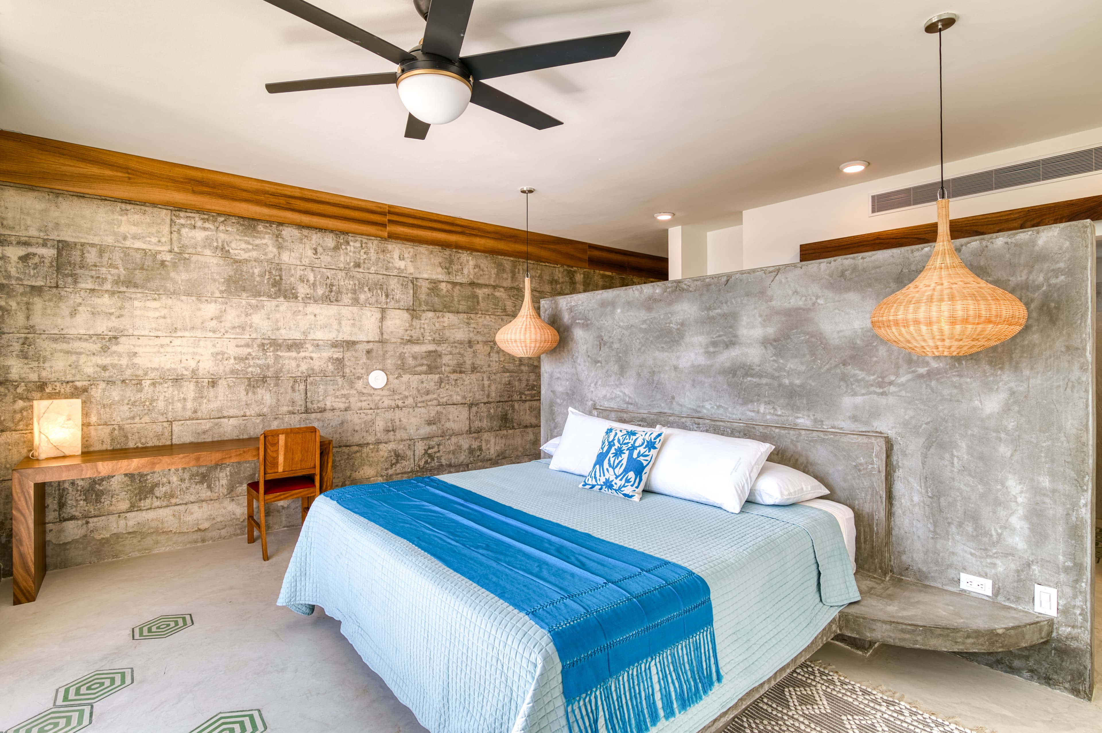 A modern bedroom featuring a concrete accent wall, a queen-sized bed with blue bedding, and stylish pendant lights.