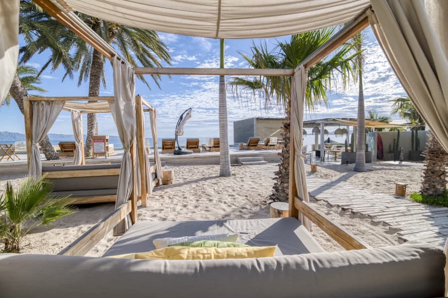 A view of a sandy beach area with cabanas, palm trees, and lounge chairs under a blue sky.