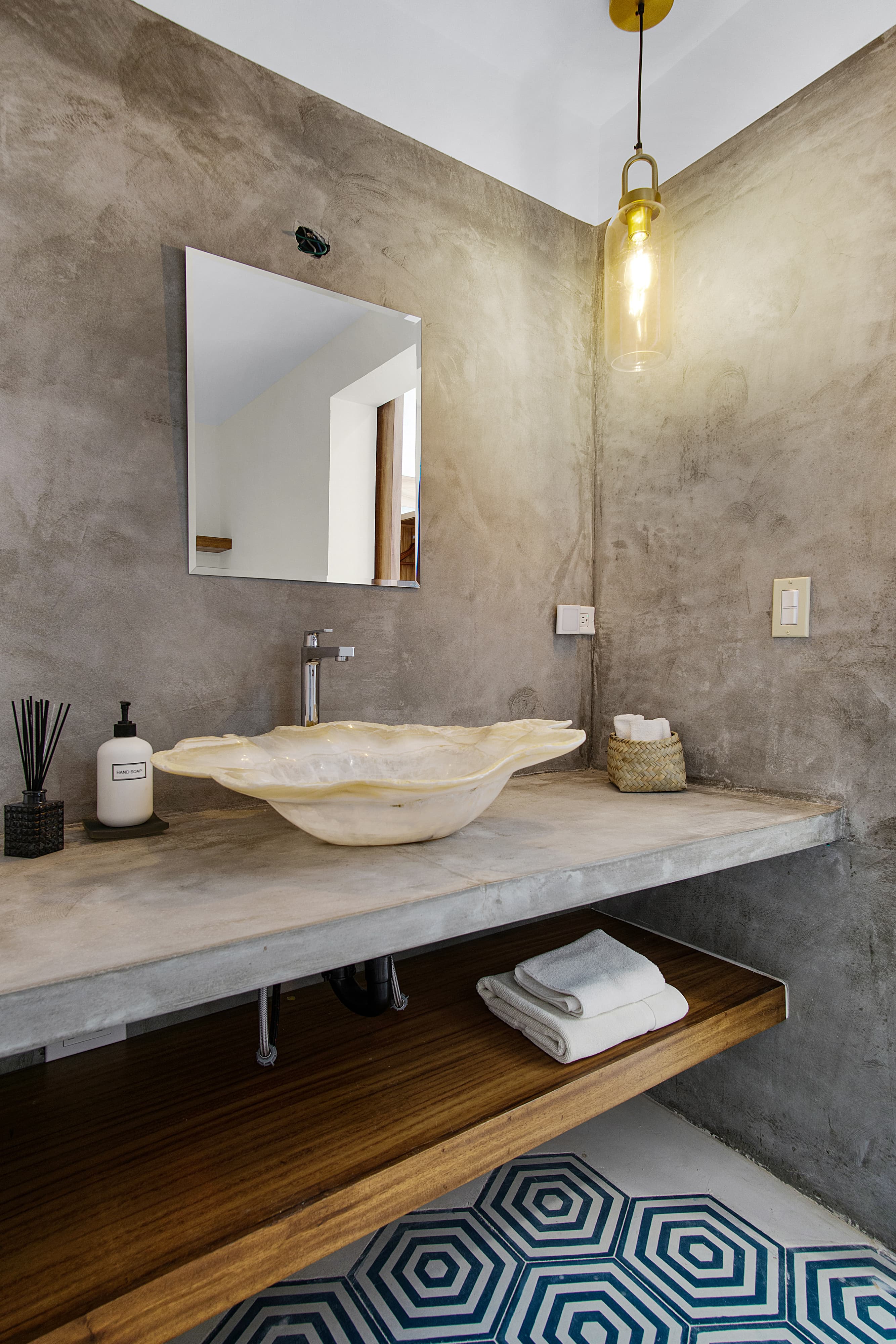 A modern bathroom featuring a concrete sink, wooden shelf, and geometric patterned rug.