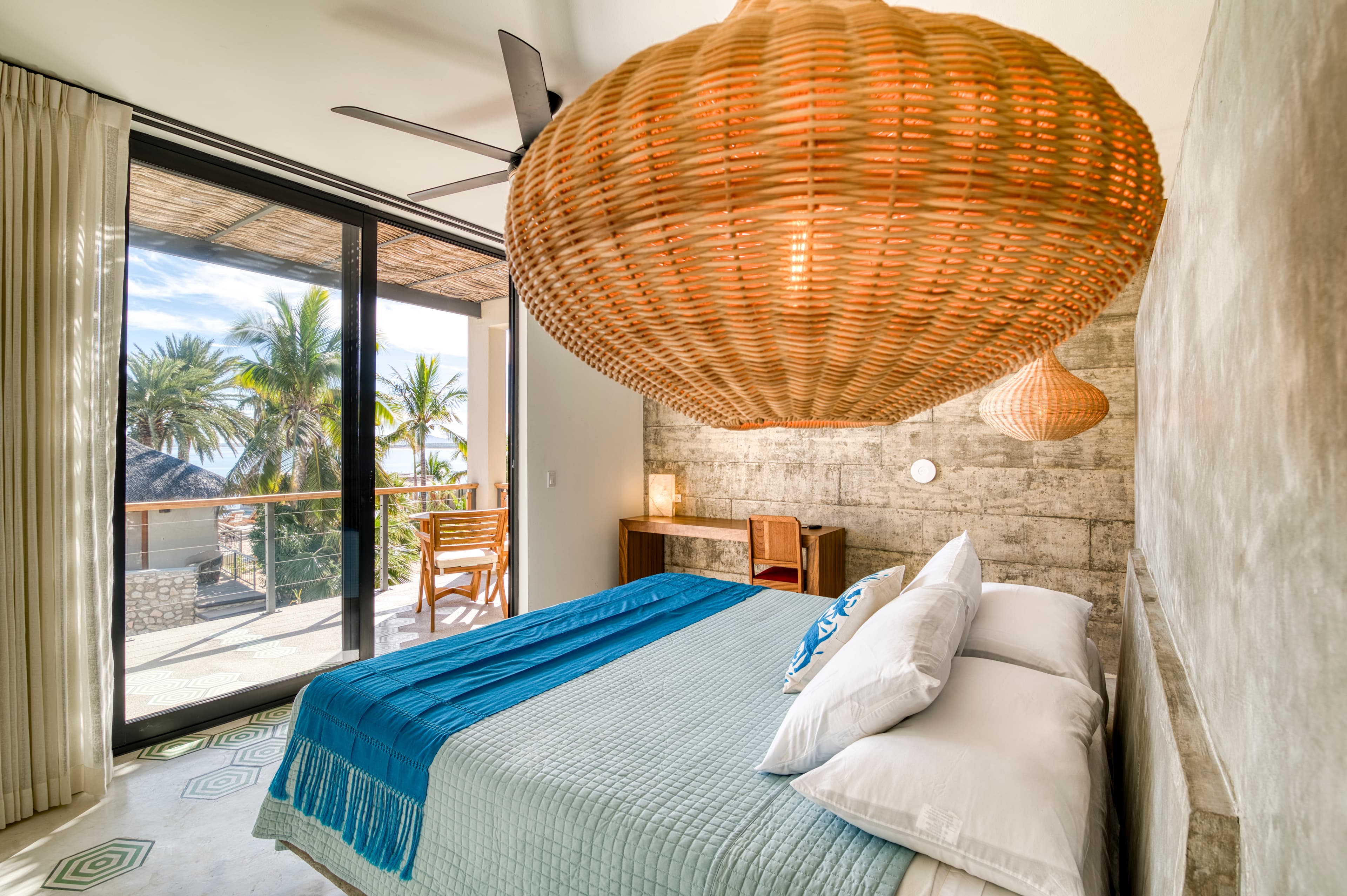 Bright and modern bedroom with a large woven light fixture, a comfortable bed, and a view of palm trees through glass doors.