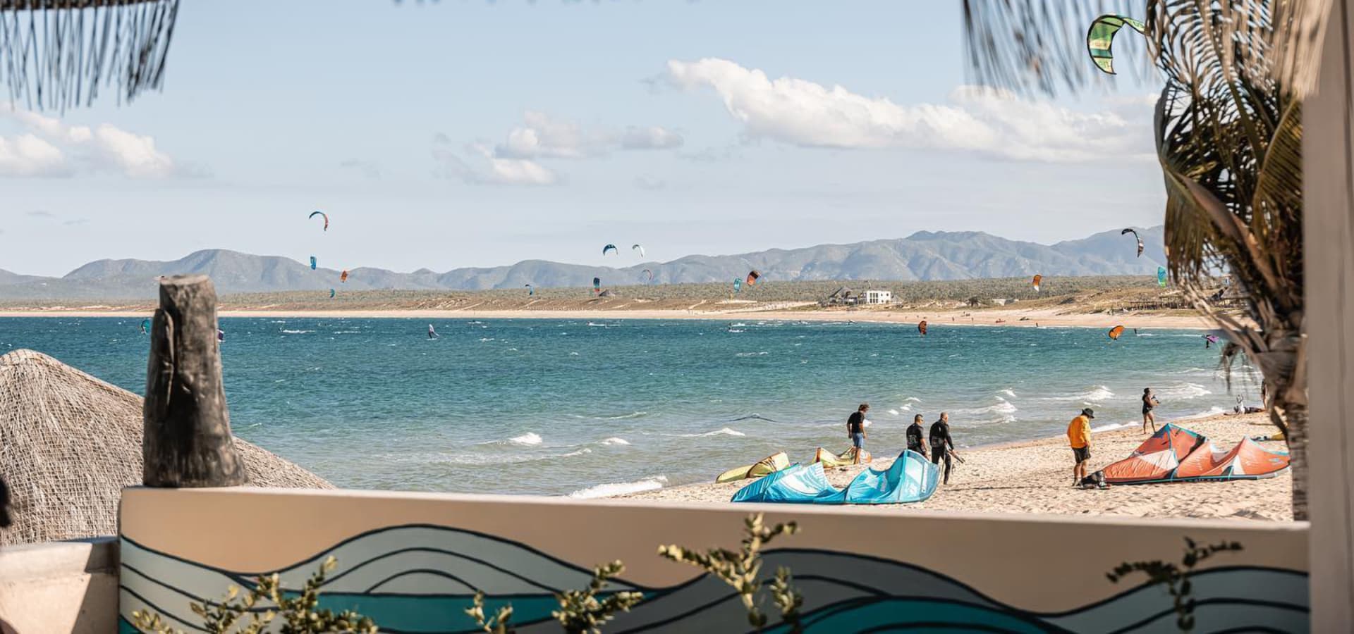 A scenic view of a beach with kite surfers and colorful kites against a backdrop of mountains and blue skies.
