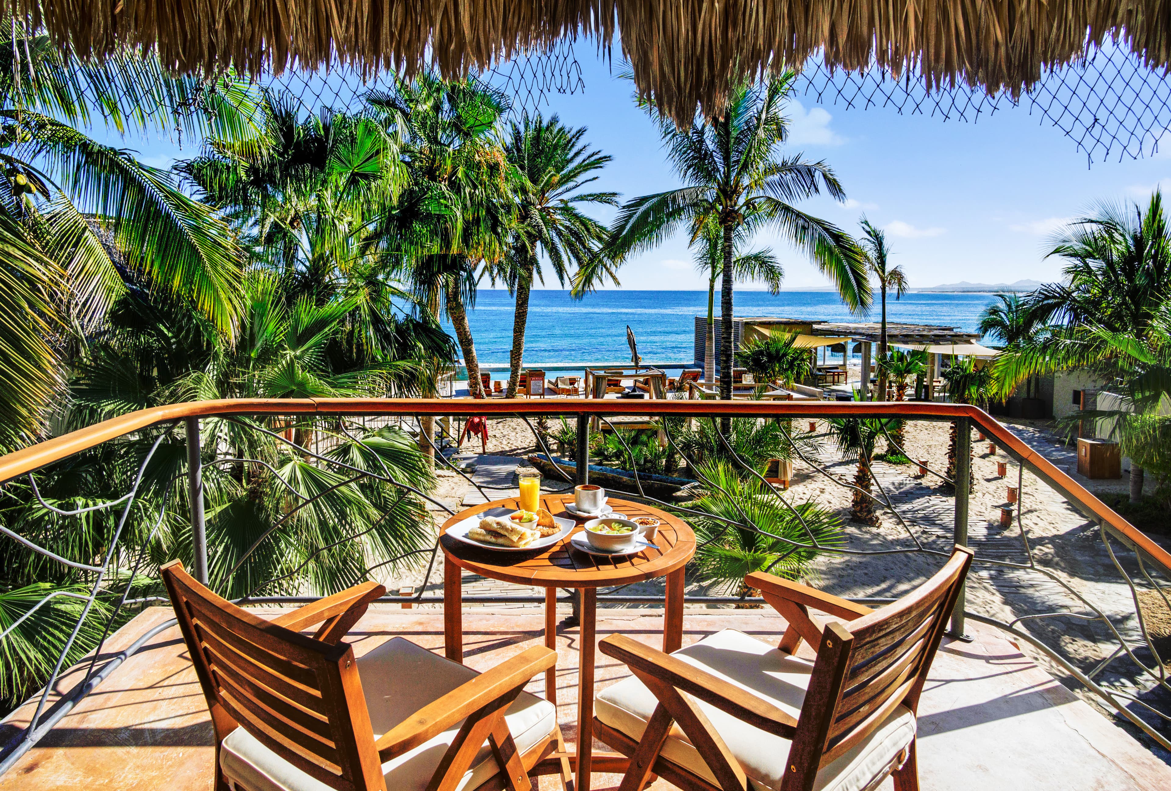A balcony with two chairs overlooks a sandy beach surrounded by palm trees and the ocean.
