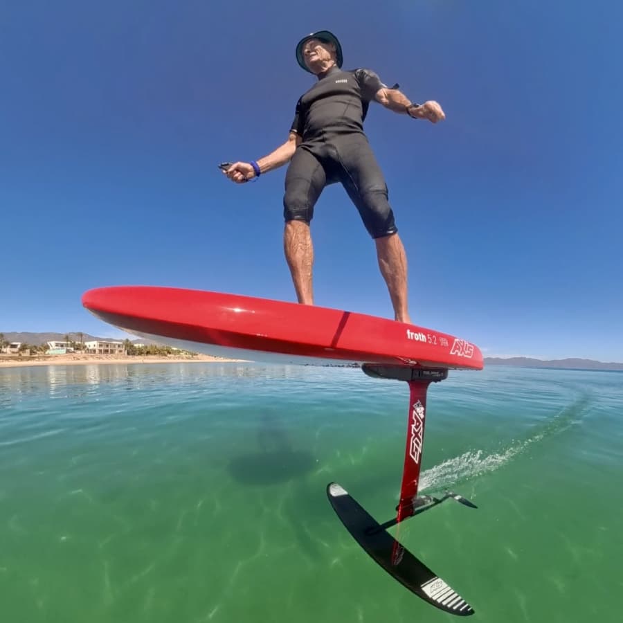 A person is hydrofoiling on a red board above clear water under a blue sky.