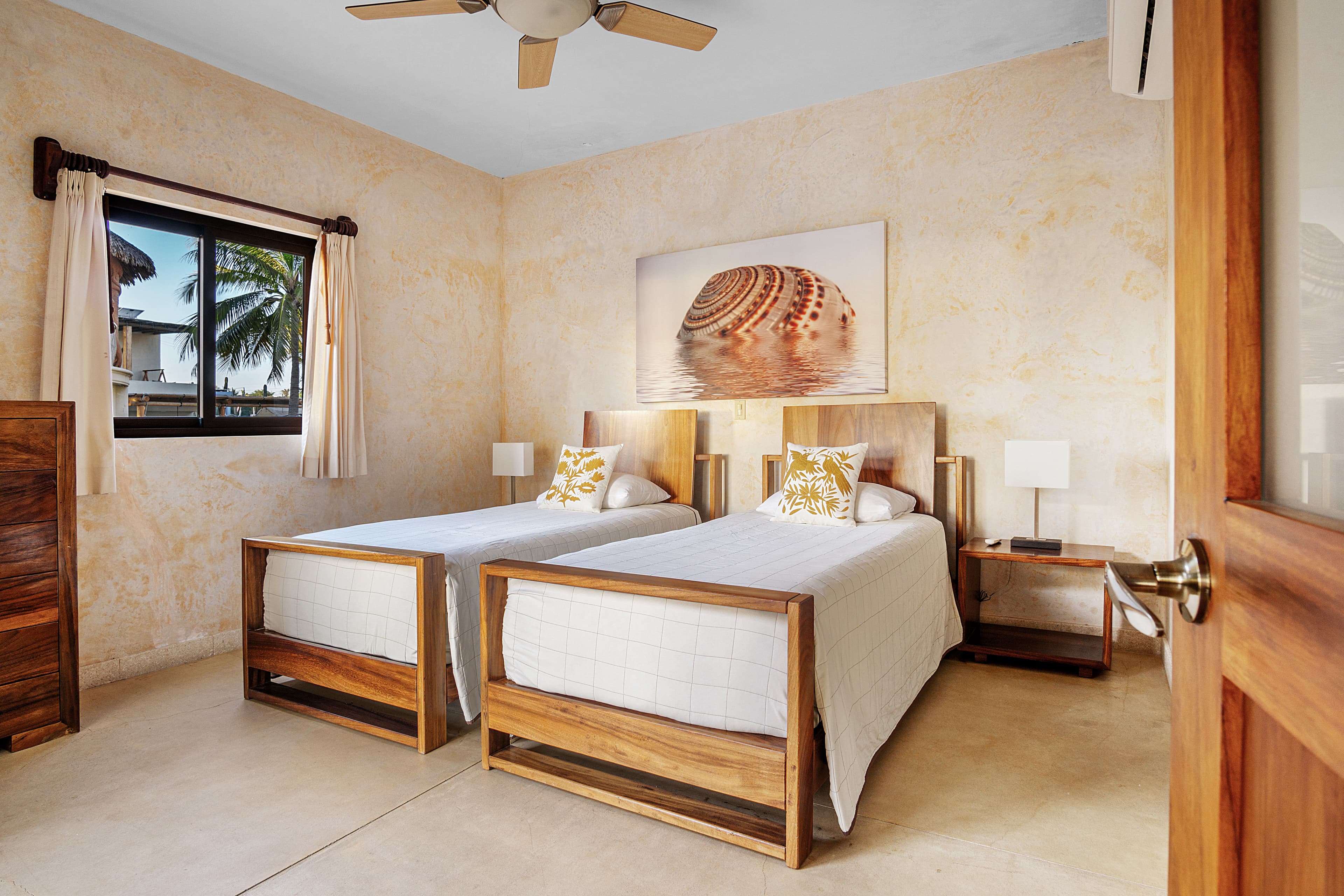 Bright bedroom featuring two wooden twin beds, decorative pillows, and a large seashell art piece on the wall.