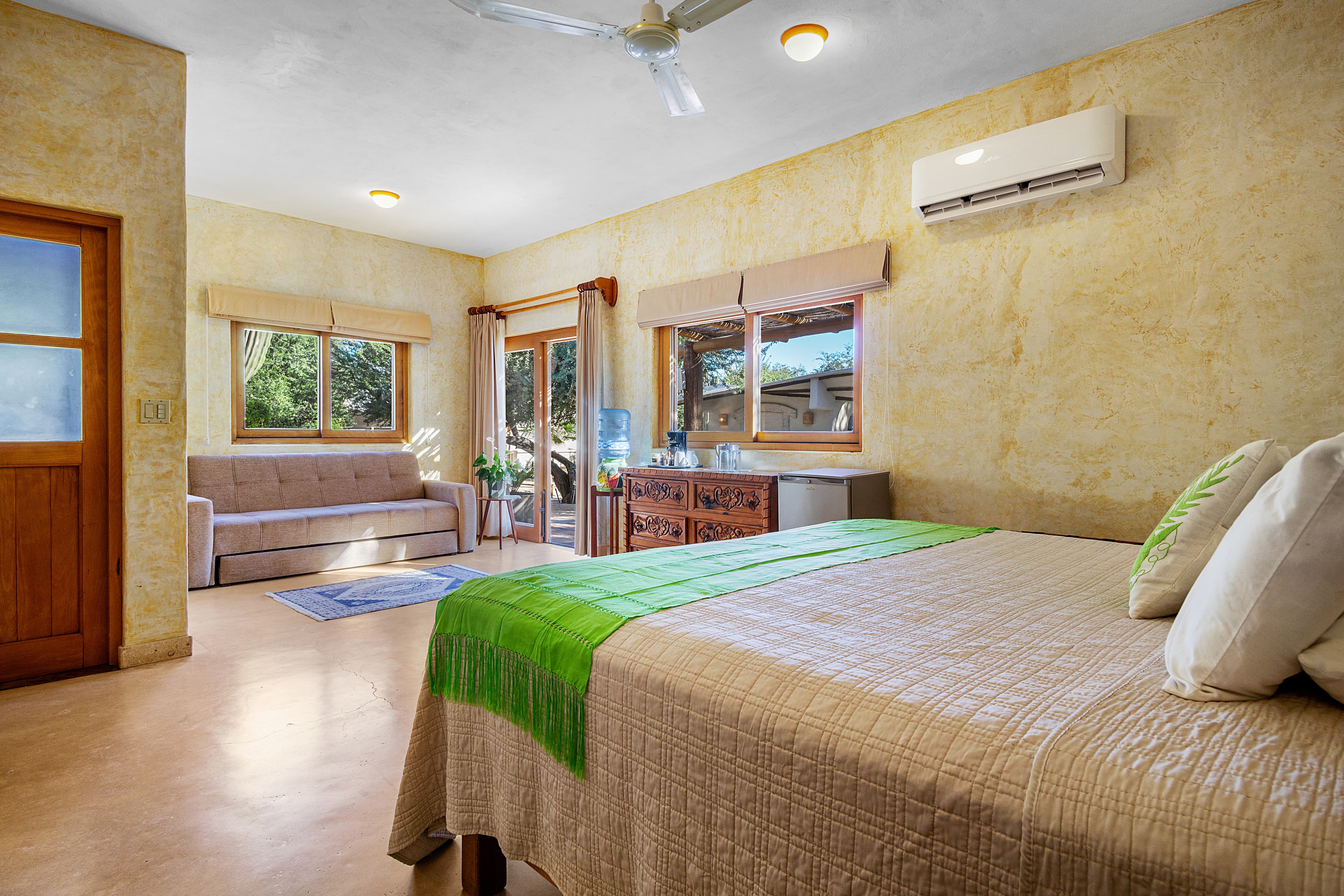 A cozy bedroom featuring a bed with green accents, a sofa, and bright windows overlooking a garden.