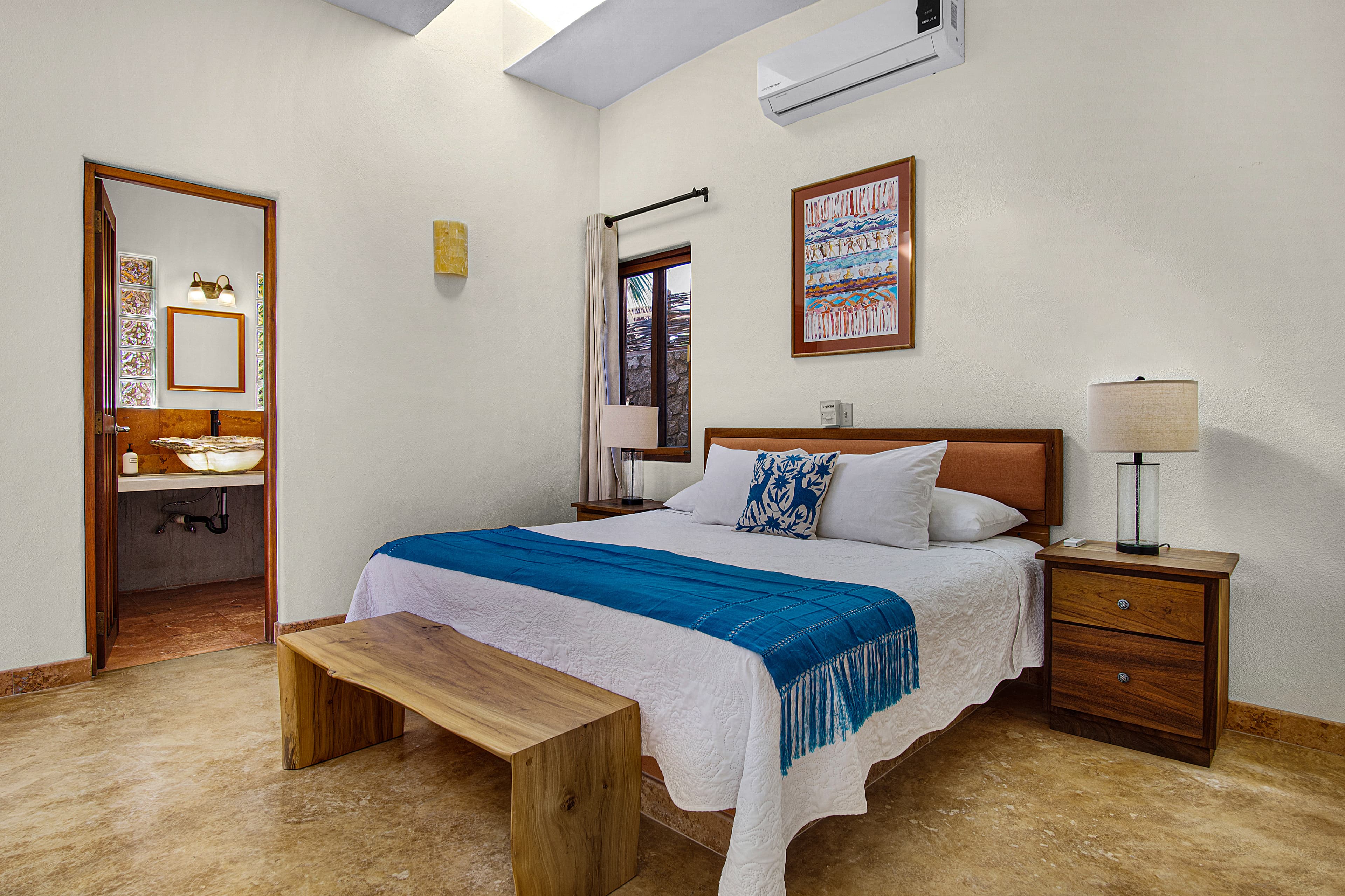 A cozy bedroom featuring a bed with blue and white bedding, wooden furniture, and an en-suite bathroom visible through a doorway.