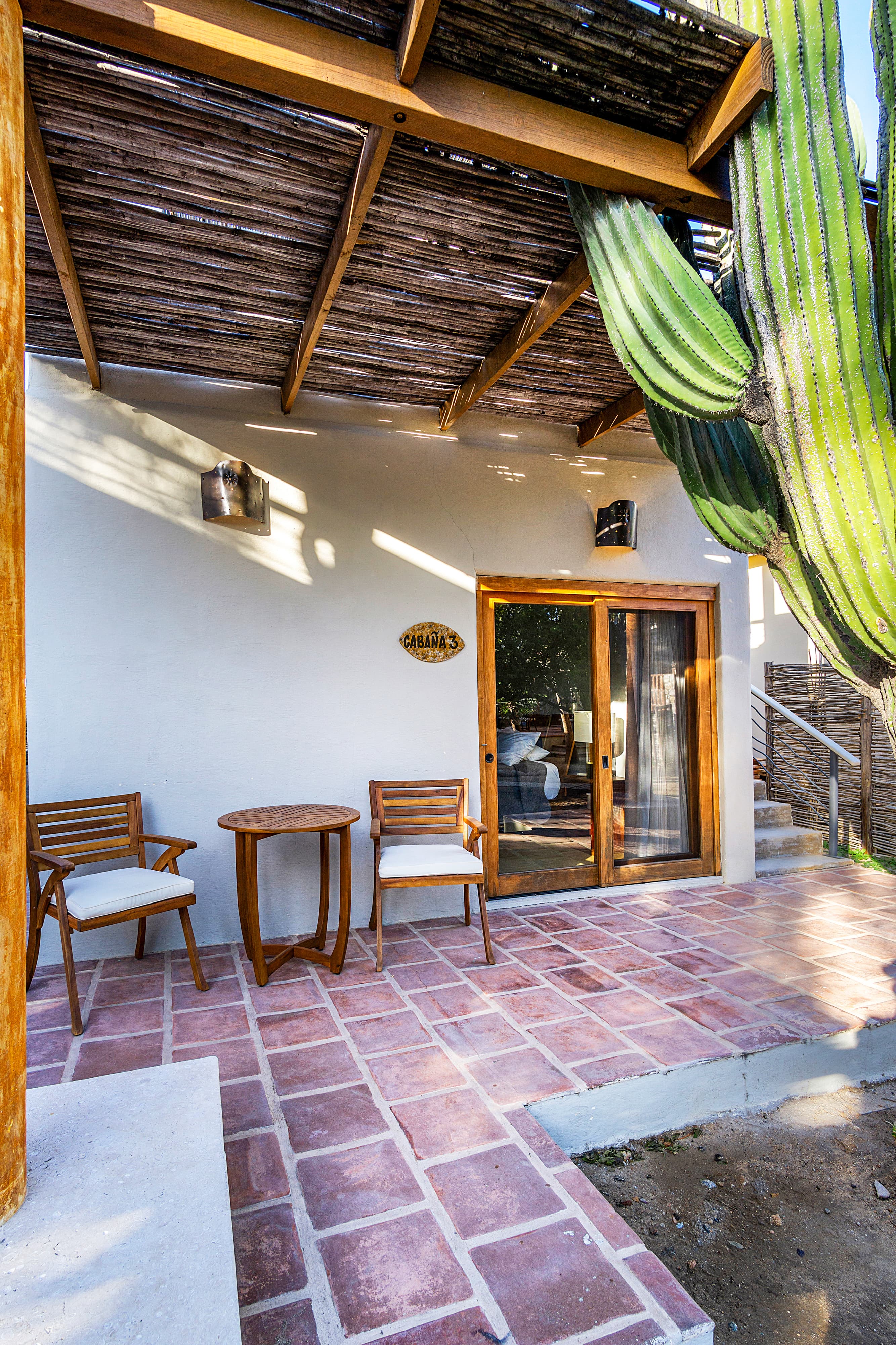 A cozy outdoor seating area with two chairs and a table, framed by a tall cactus and a partially shaded porch.