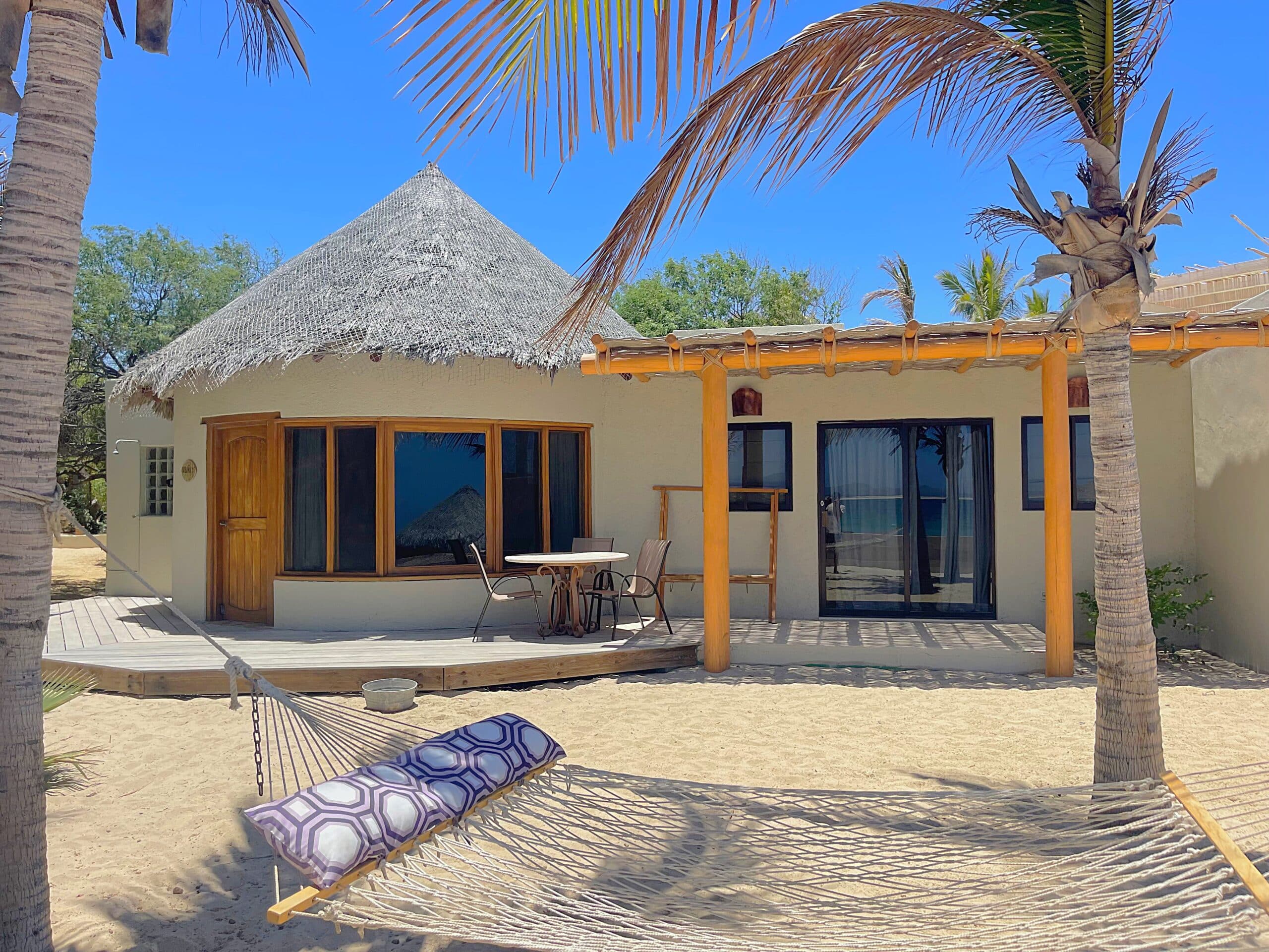 A cozy beachside bungalow with a thatched roof, palm trees, and a hammock in the sand.