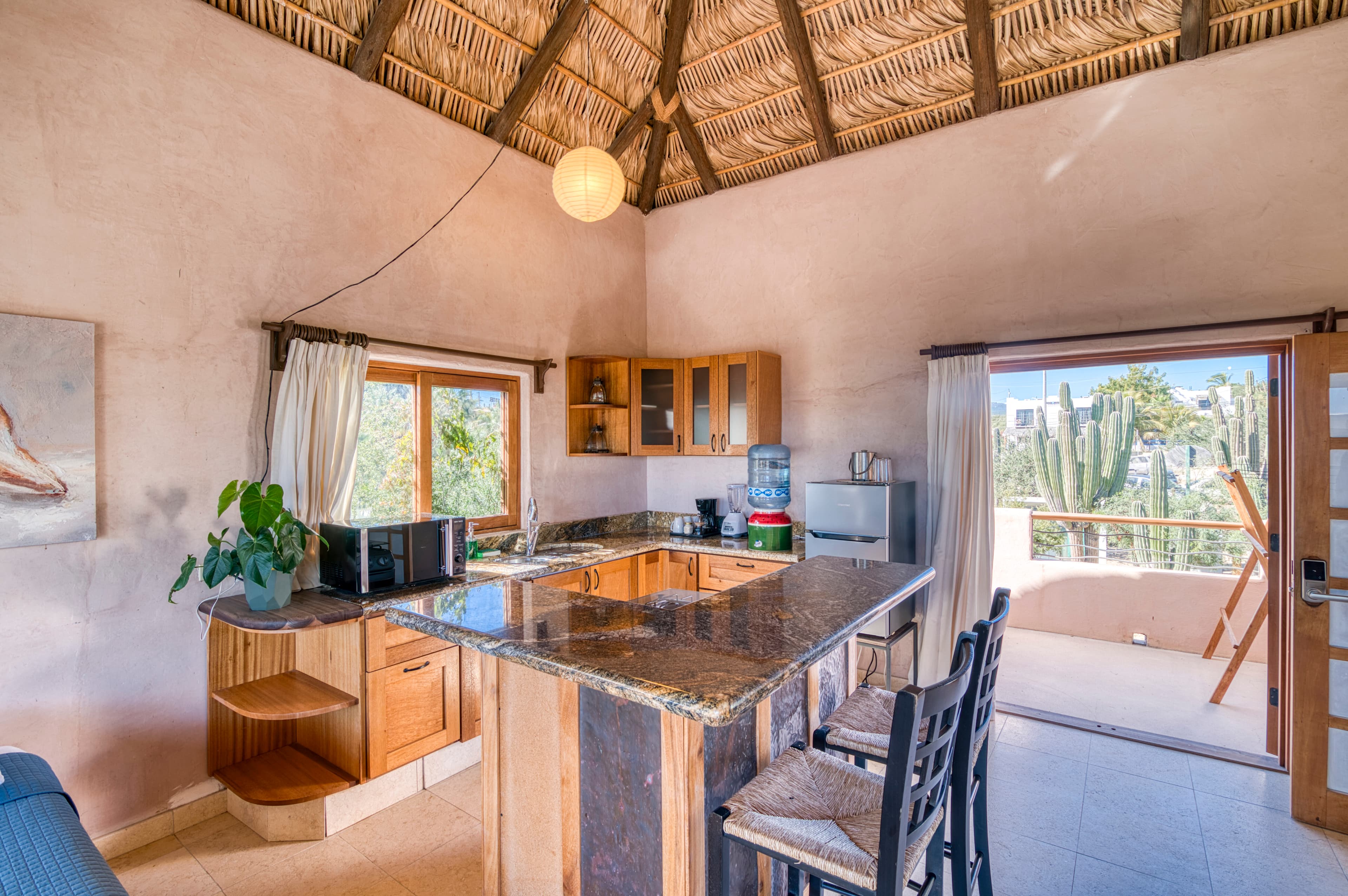 A cozy kitchen with a high thatched ceiling, wooden cabinets, and a scenic view through the open door.