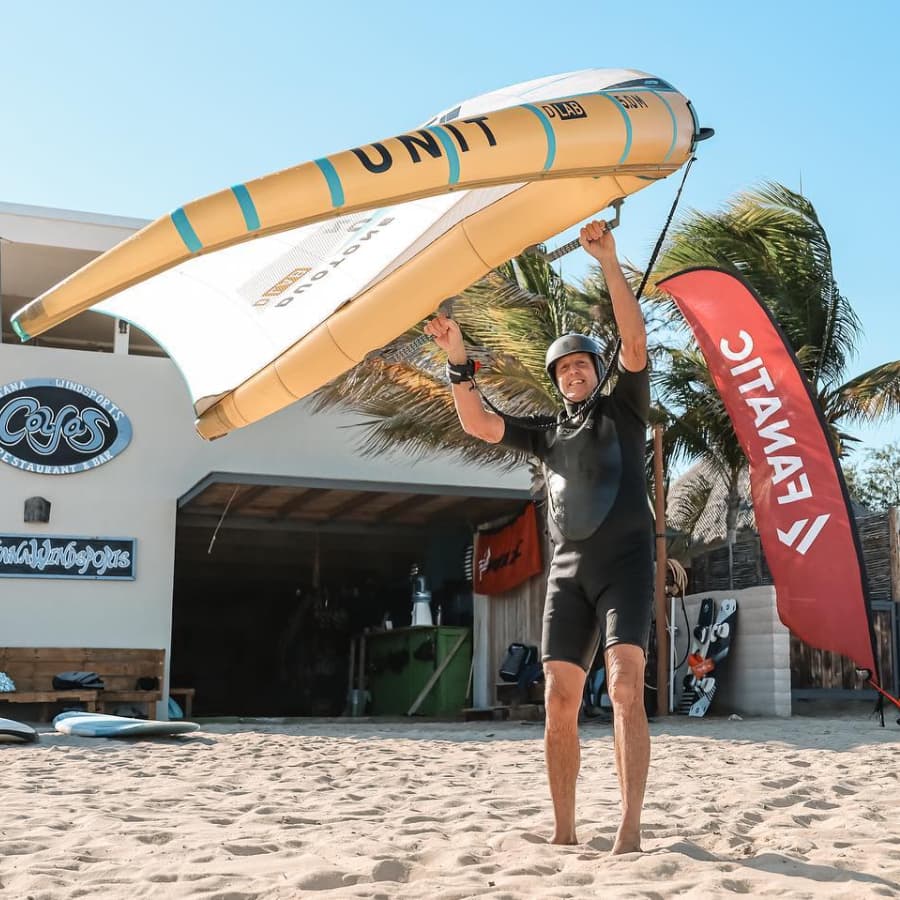 A man in a wetsuit joyfully holds up a windsurfing sail on a sandy beach.