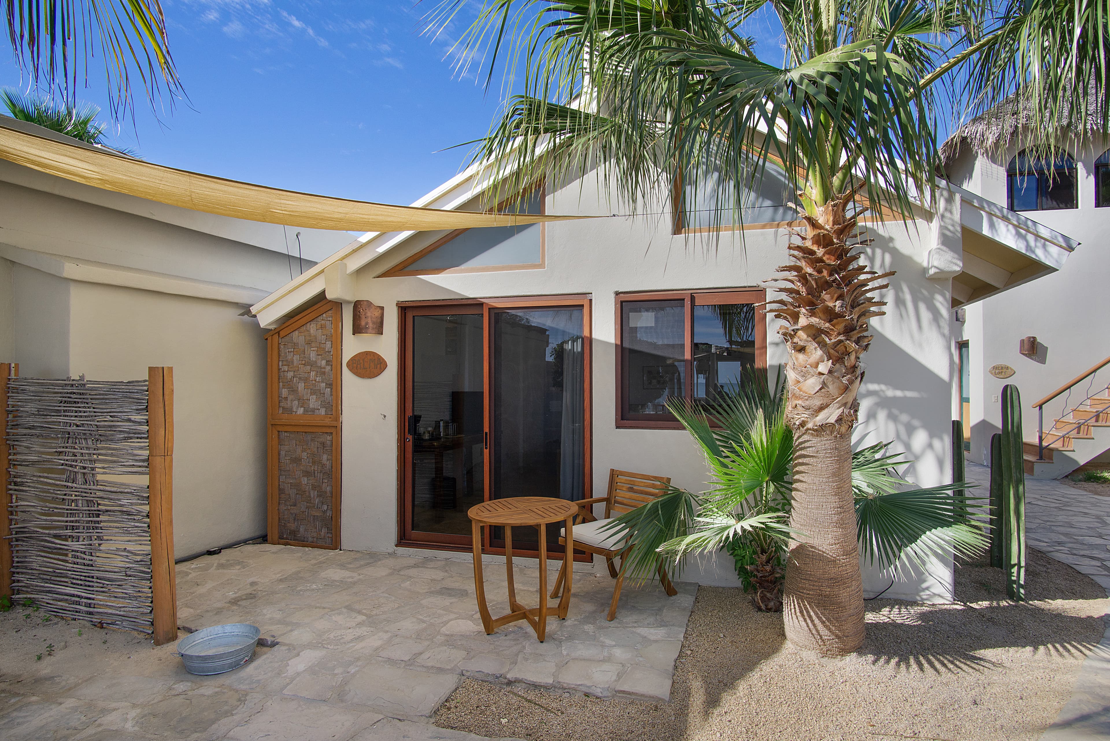 Exterior view of a cozy house surrounded by palm trees and a small seating area on a stone patio.