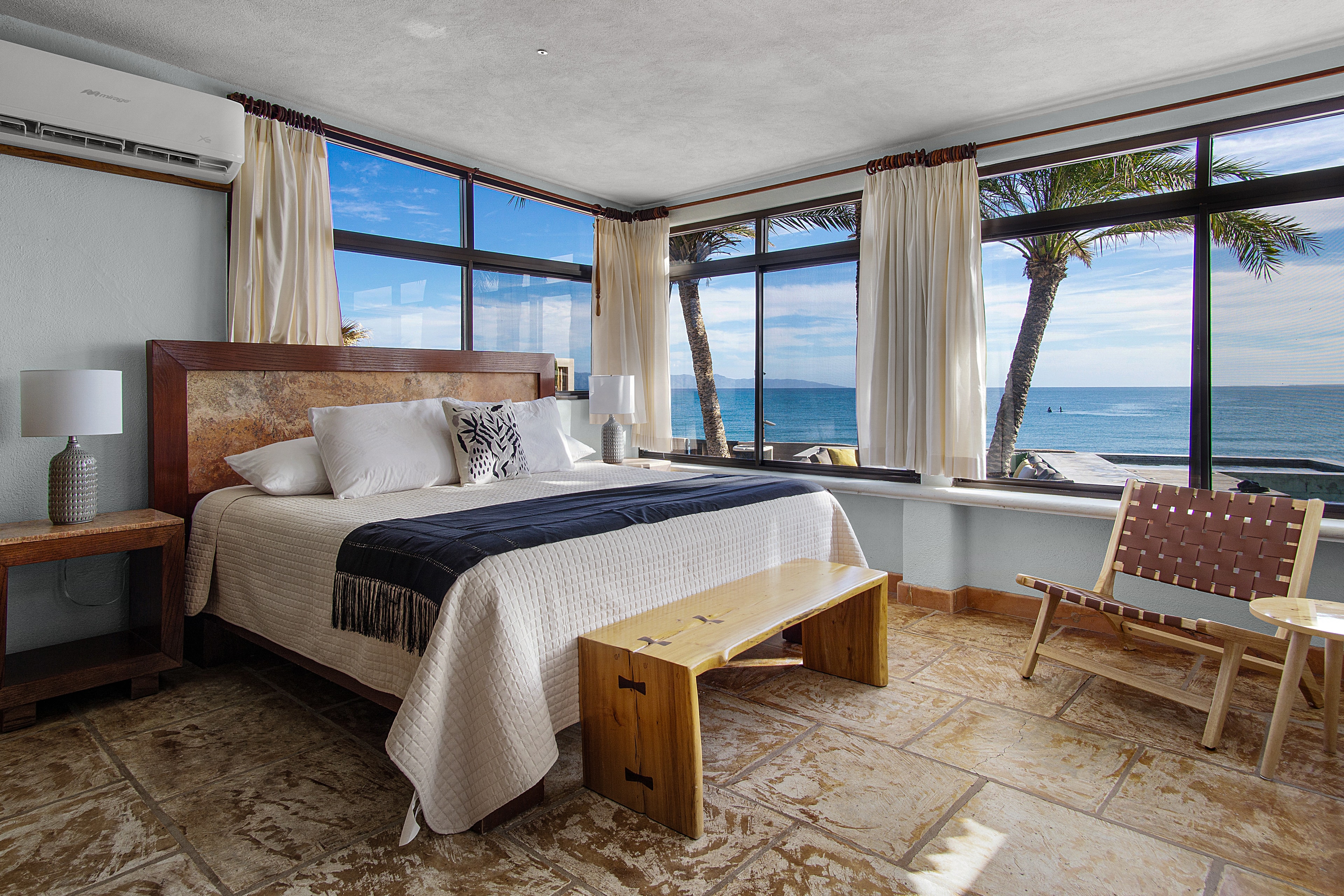 A modern bedroom with large windows showcasing a beach view, featuring a bed, wooden furniture, and palm trees outside.
