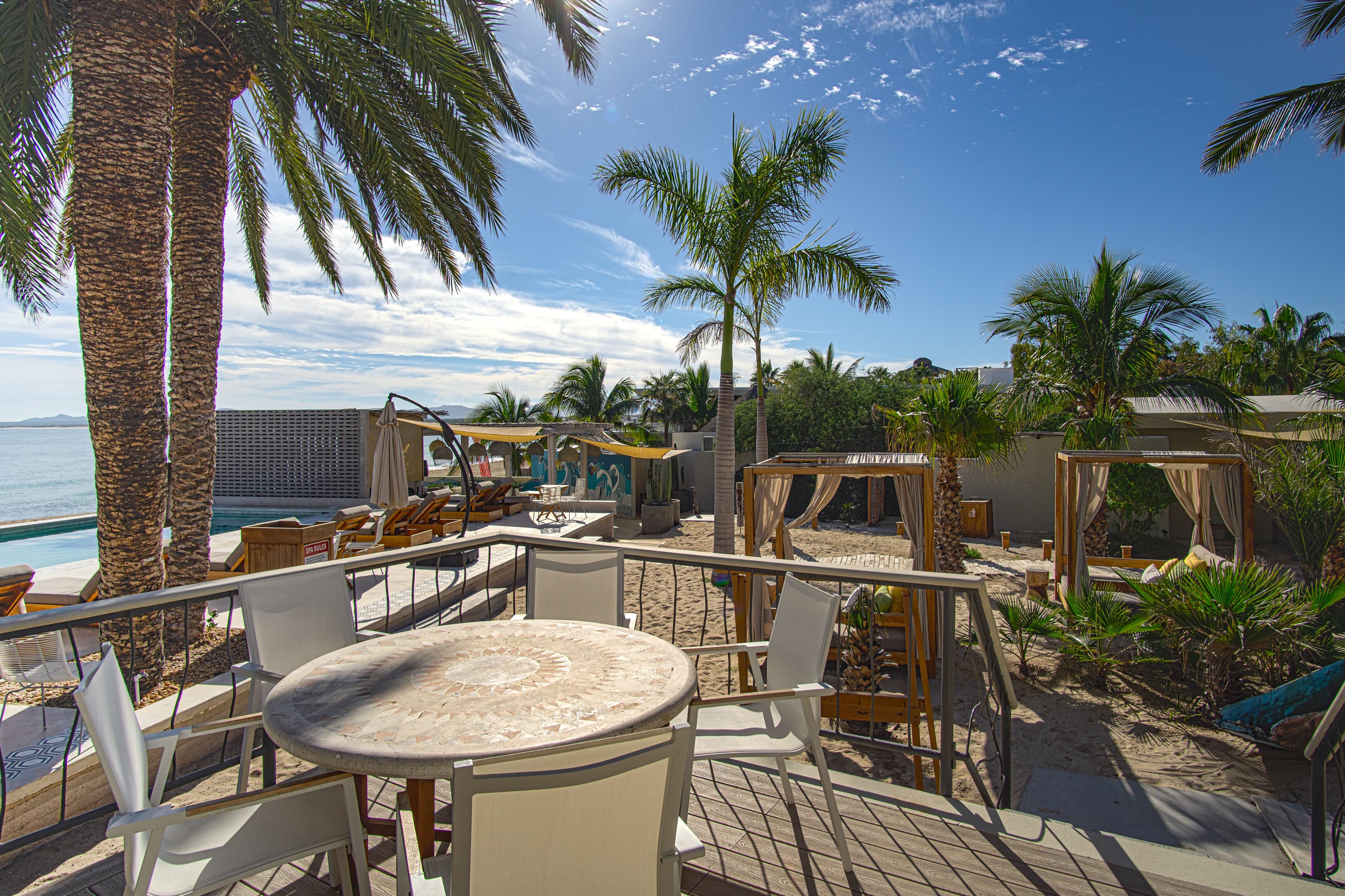 A sunny seaside area featuring palm trees, lounge chairs, and a table on a wooden deck.