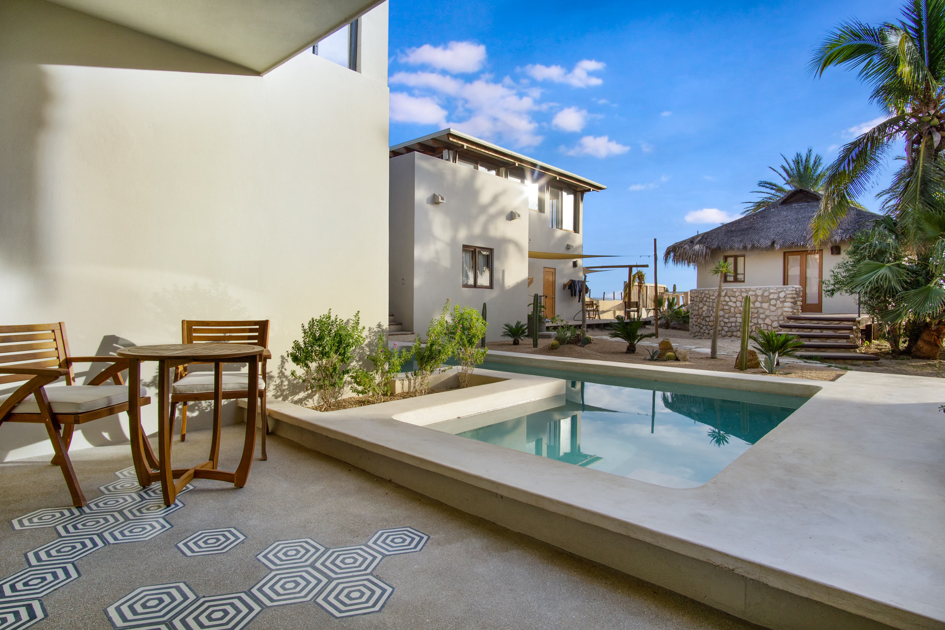 A serene outdoor space features a small pool surrounded by plants and a seating area near two modern buildings.