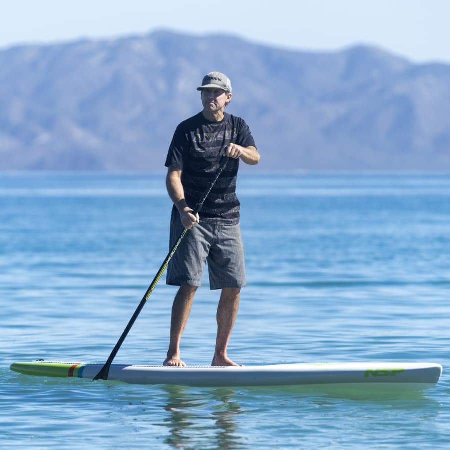 A man stands on a paddleboard in calm water with mountains in the background.