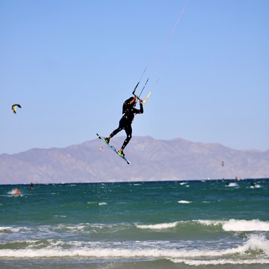 A kitesurfer performs a jump above the ocean with mountains in the background.